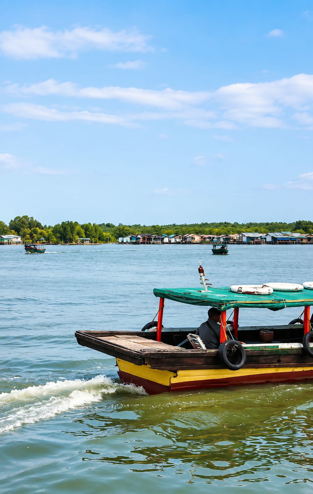 In the city of Medan, Indonesia, a traditional wooden boat with a yellow and red hull, powered by an unseen engine, is creating a wake as it moves through calm, greenish water. The boat is covered by a green canopy supported by red poles, and a person wearing a dark shirt sits at the helm. Two spare tires are attached to the sides of the boat, and a life preserver rests on the canopy. In the background, a line of stilted houses with colorful roofs forms a village along the tree-lined shore. Another, smaller green boat is visible in the distance. The sky above is a clear blue with scattered white clouds.
