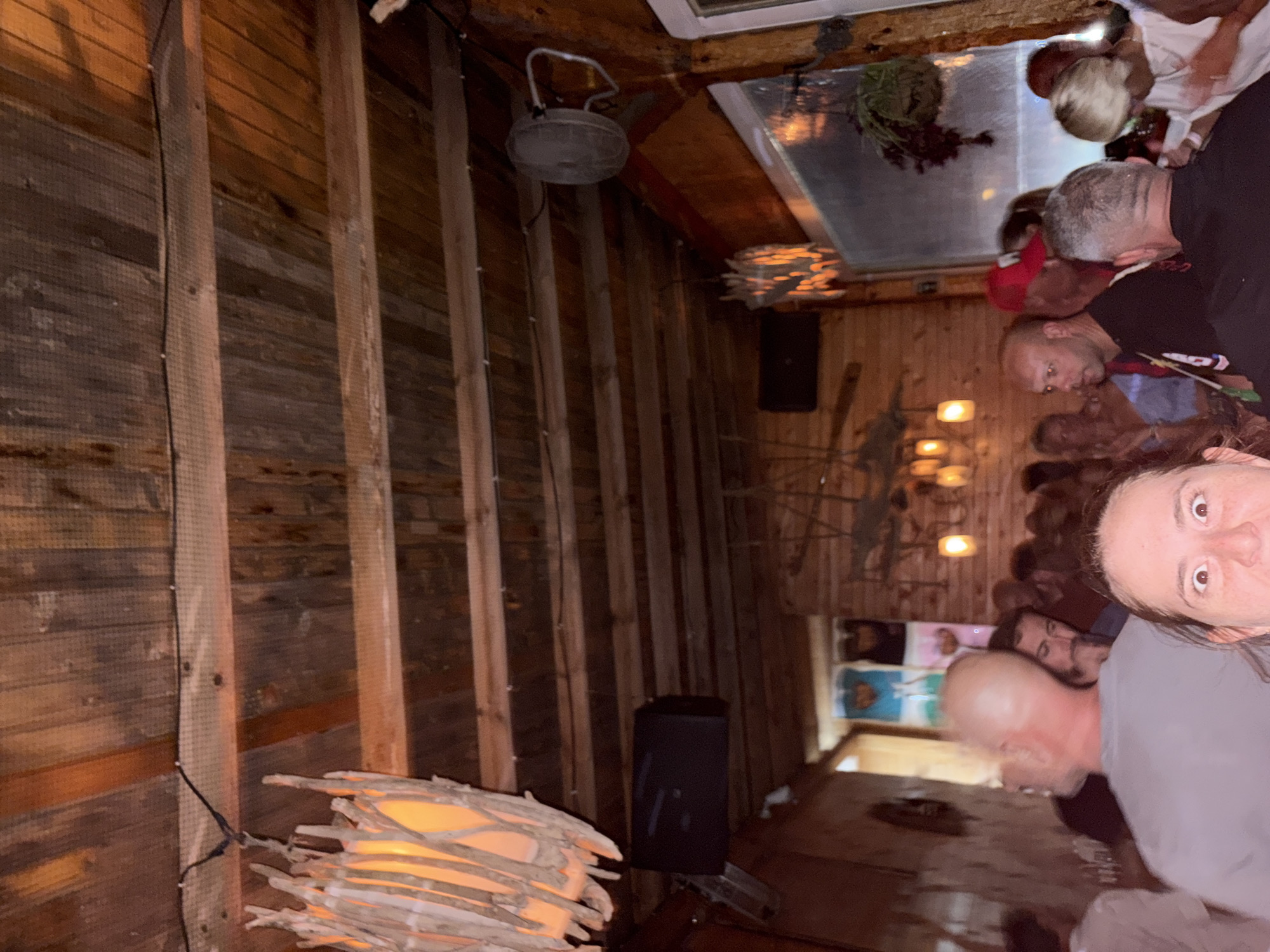 The image depicts an indoor scene, likely a bar or restaurant, with a rustic wooden interior. The ceiling is constructed of wooden planks. There is a prominent decorative light fixture made of branches to the left of the frame, emitting a warm glow. A speaker is mounted on the wall below the light fixture. Further into the scene, another decorative light fixture with visible bulbs is visible.
A crowd of people is present, with many faces partially visible and some in motion. A person in the foreground is looking directly at the camera with a surprised expression, their eyes wide open. To the left, a person with a bald head is seen from behind, wearing a grey t-shirt. Other individuals are visible in the background, some interacting and others appearing to be observing. The overall lighting is dim, with warm tones from the decorative lights. The setting appears to be a social gathering. The location is identified as Lozenets, Bulgaria.