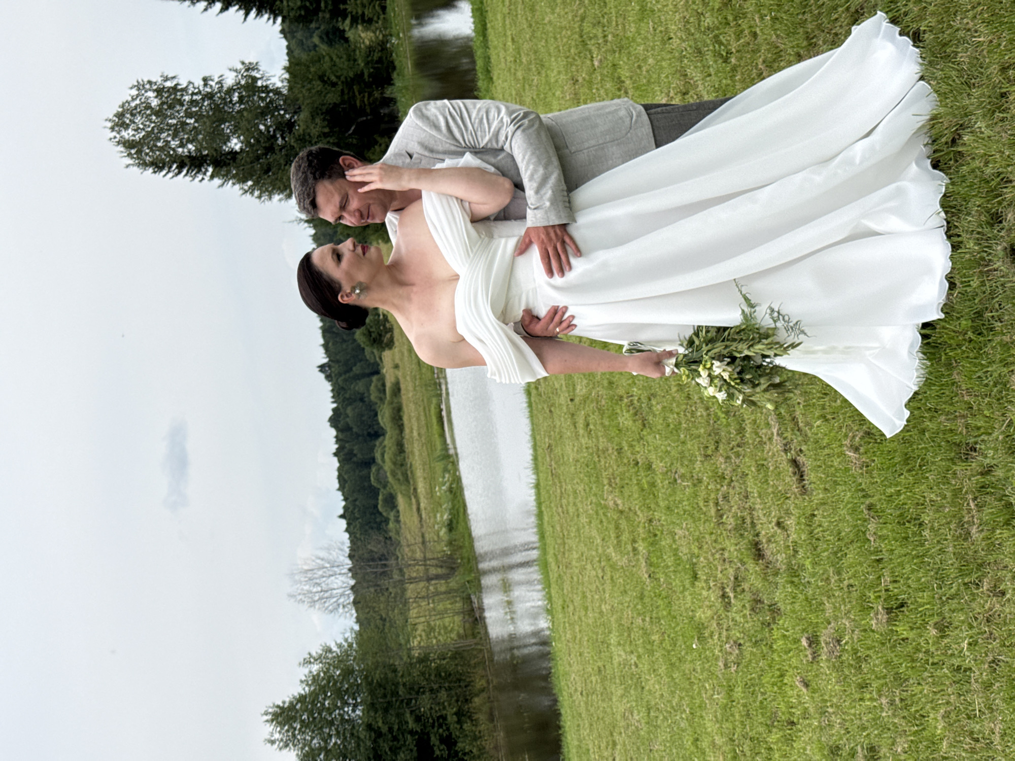A couple, a man and a woman, are standing in a grassy field next to a body of water. The woman is wearing a white off-the-shoulder wedding dress and holding a bouquet of flowers. The man is wearing a light-colored suit jacket and pants and has his arm around the woman. They are looking at each other. The background shows trees and a cloudy sky. The location is identified as Svetloye, Russia, though specific landmarks are not visible.