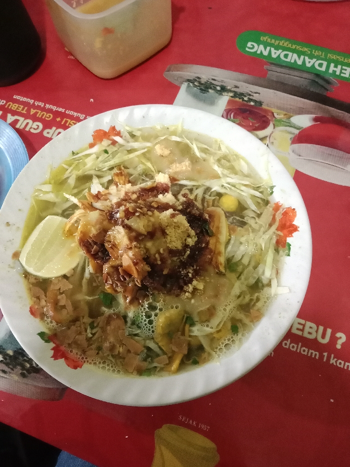 A white bowl containing a yellow broth soup with shredded cabbage, fried noodles, and a dark brown sauce is placed on a red table, likely in a restaurant in Tugu, Indonesia, with coordinates -6.975, 110.332. The soup is topped with a slice of lemon and a few crispy noodles, along with some fried white food, which may be tofu. The table is adorned with red and white checkered paper. In the background, a partially visible plastic container with a yellow liquid sits on the table. To the right of the soup bowl, a red and green sign with the words "Eh Dandang" and "rasasi teh seanggunya" can be seen. The sign is likely advertising a beverage, suggesting that the photo was taken at a food stall or restaurant. The image is taken from an overhead perspective, likely by a customer dining at the establishment.