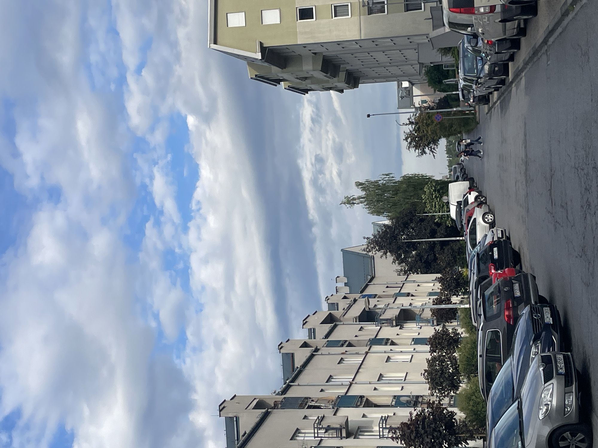 The image shows a street scene in an urban area, with buildings lining both sides of the road. Several cars are parked along the roadside. In the distance, people are visible walking on the street. The sky is filled with clouds, and the buildings appear to be residential apartments.
