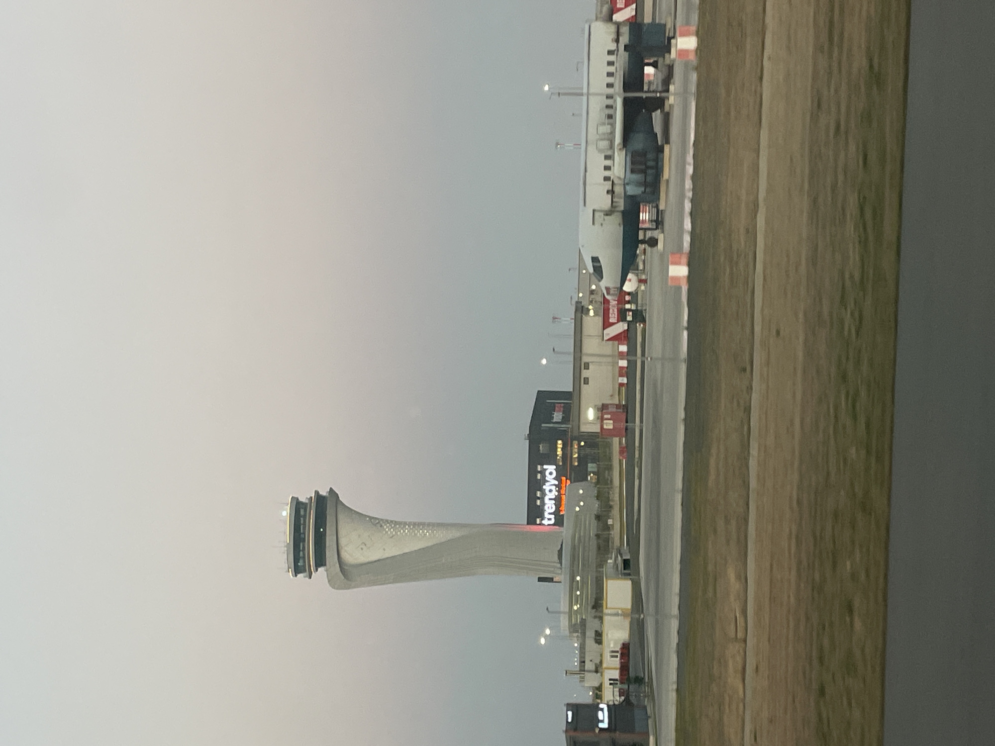 In Beykoz, Turkey, the image captures the Istanbul Airport's iconic control tower, a slender, modern structure with a bulbous top housing the control room. The tower stands tall against a pale, overcast sky, its white exterior adorned with subtle patterned detailing. At the base of the tower, a dark building displays a bright red "trendyol" logo. To the right, a decommissioned aircraft, painted white and blue, rests on the tarmac. Red and white striped barriers delineate the airport's operational areas. The foreground shows a strip of dry, brown grass and a dark gray surface, possibly a road or a runway edge. The overall scene is quiet and functional, indicative of an airport environment.