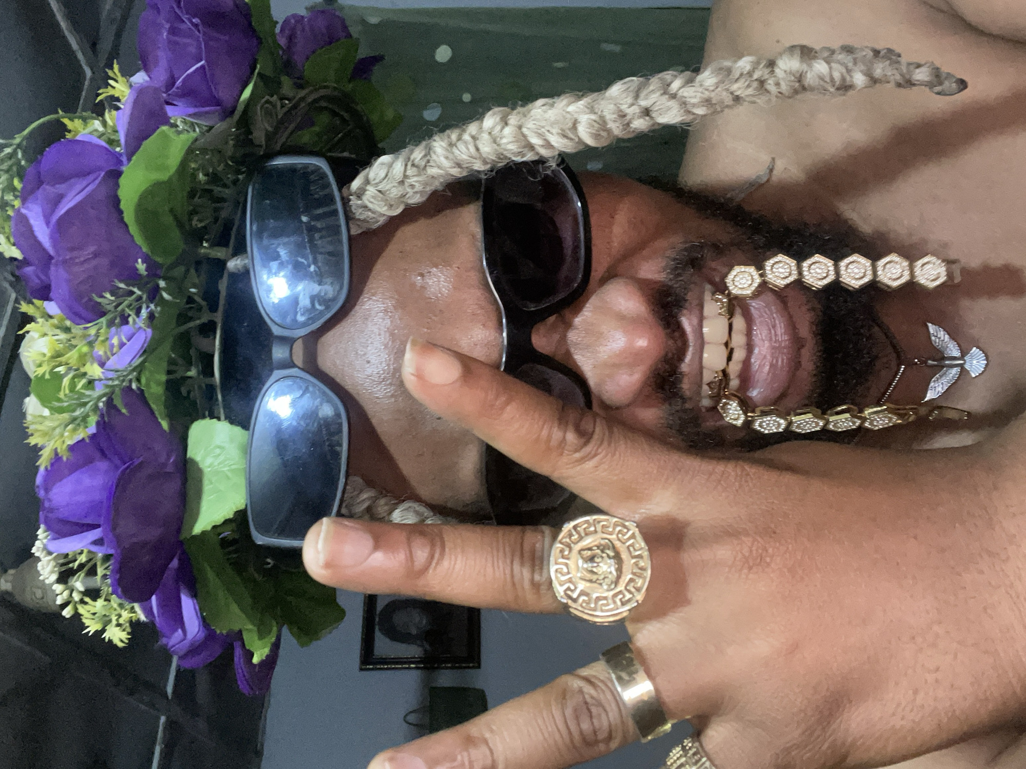 The image depicts a man with dreadlocks and a beard, smiling with his tongue visible. He is wearing two pairs of sunglasses, one on top of the other, and a crown of purple flowers. He is also wearing a gold chain on his beard and rings on his fingers, including one with a Versace-like Medusa head design. He is giving the peace sign with his fingers. The background is blurry, but appears to be an indoor setting. There is no clear evidence to suggest that the location is Jos, Nigeria.