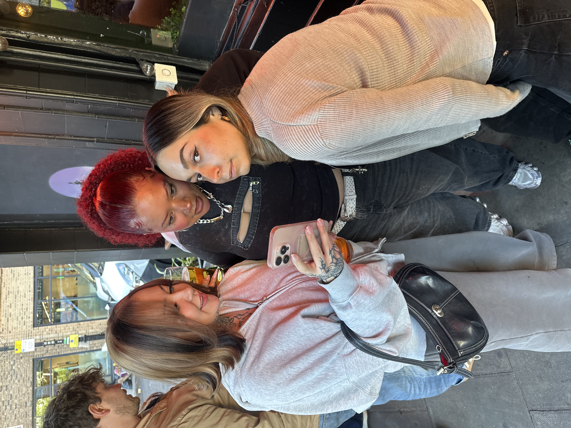Three young women are captured at an outdoor establishment in what appears to be London, UK. The architecture indicates an urban setting. The woman at the bottom of the frame is holding a phone and taking a selfie. She is wearing a grey hoodie. Next to her is a small beer. The woman next to her is wearing a black top and has red hair. The third woman is wearing a striped beige sweater. She is leaning her face onto the black-topped woman. A man is sitting to the bottom left of the frame. He is wearing a brown jacket.