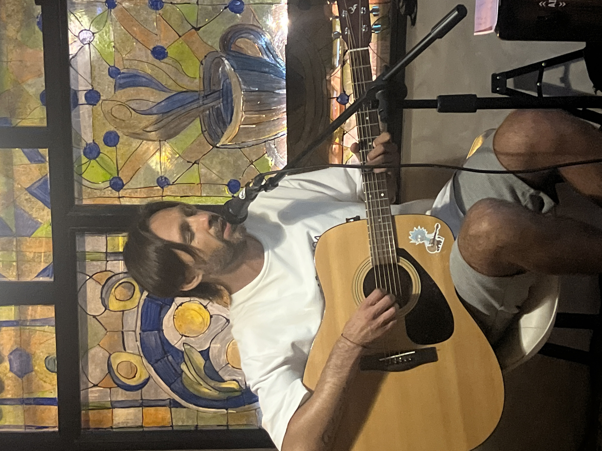 A man is seated on a white stool, playing an acoustic guitar and singing into a microphone in front of a stained-glass window. He is wearing a white t-shirt and gray shorts. The stained-glass window features abstract designs with blue, yellow, and green colors, including a depiction of a coffee cup with steam. A microphone stand is positioned in front of him, and part of a speaker is visible to the right. The man has dark hair and a beard.