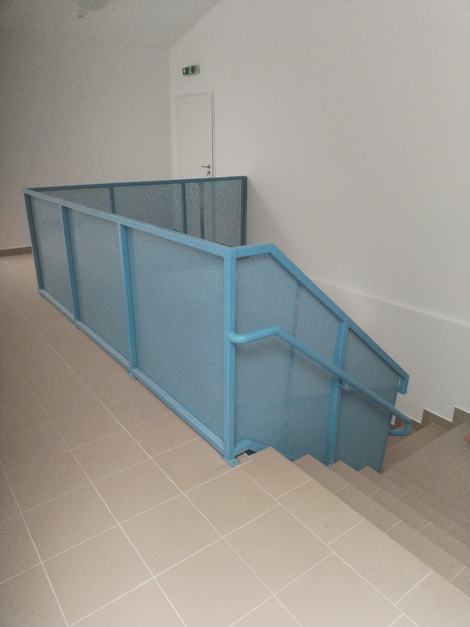 The image depicts an indoor stairway with light beige tiled flooring and a light blue metal railing. The railing is a prominent feature, enclosing the stairs and a portion of the landing. The walls are painted a light, neutral color, and an illuminated green exit sign is visible near a white door at the top of the stairs. The scene appears to be in a public building, possibly an office or apartment complex, in Bucharest, Romania. There are no people or discernible actions captured in the image. The lighting is even, suggesting an interior space with artificial illumination.