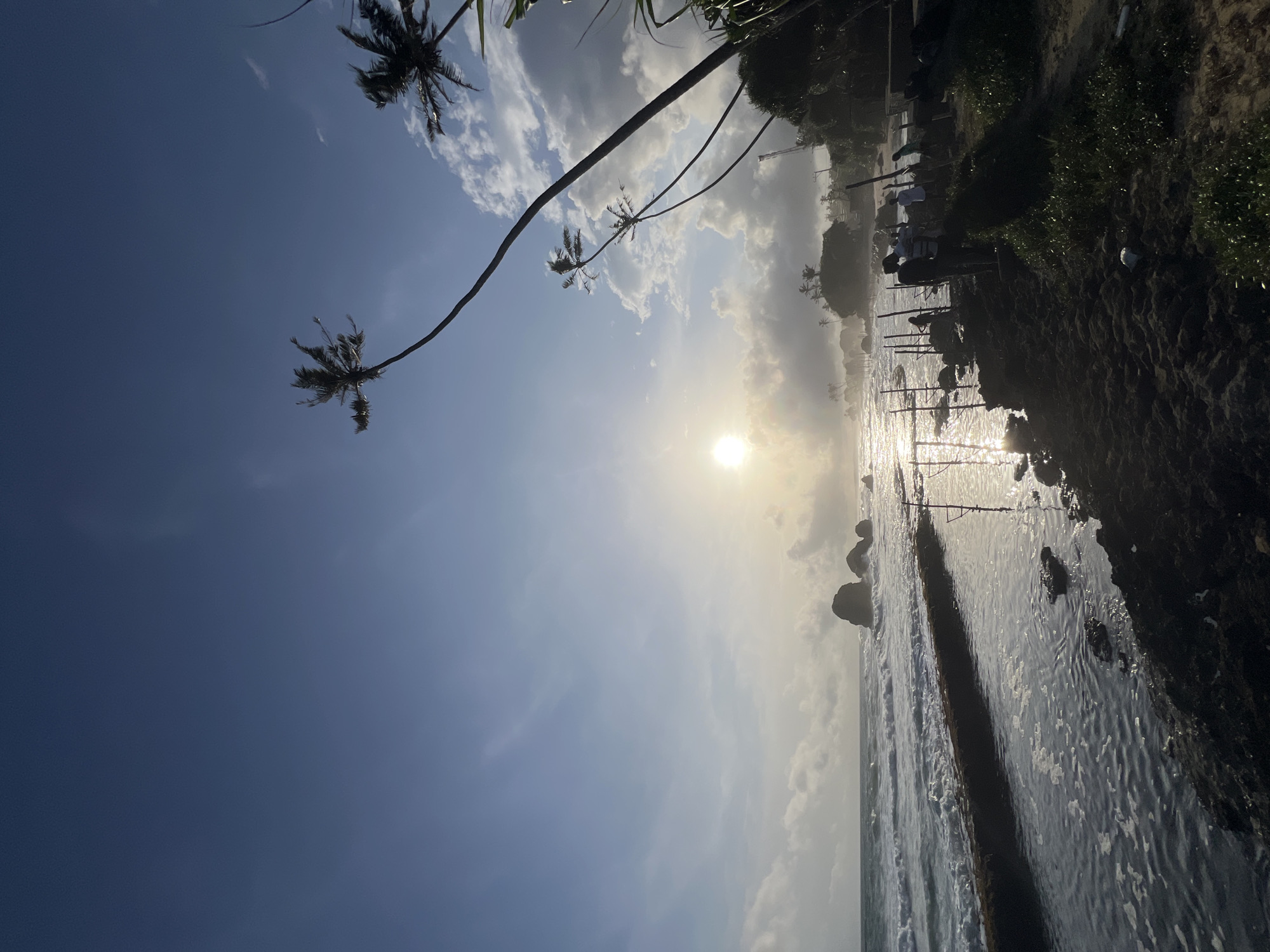 The image depicts a coastal scene in Habaraduwa, Sri Lanka. The sun shines brightly, reflecting on the water's surface. Palm trees are visible, some leaning towards the water. A shoreline is present, with vegetation and what appears to be some structures or fishing equipment on the land. The sky is partly cloudy.