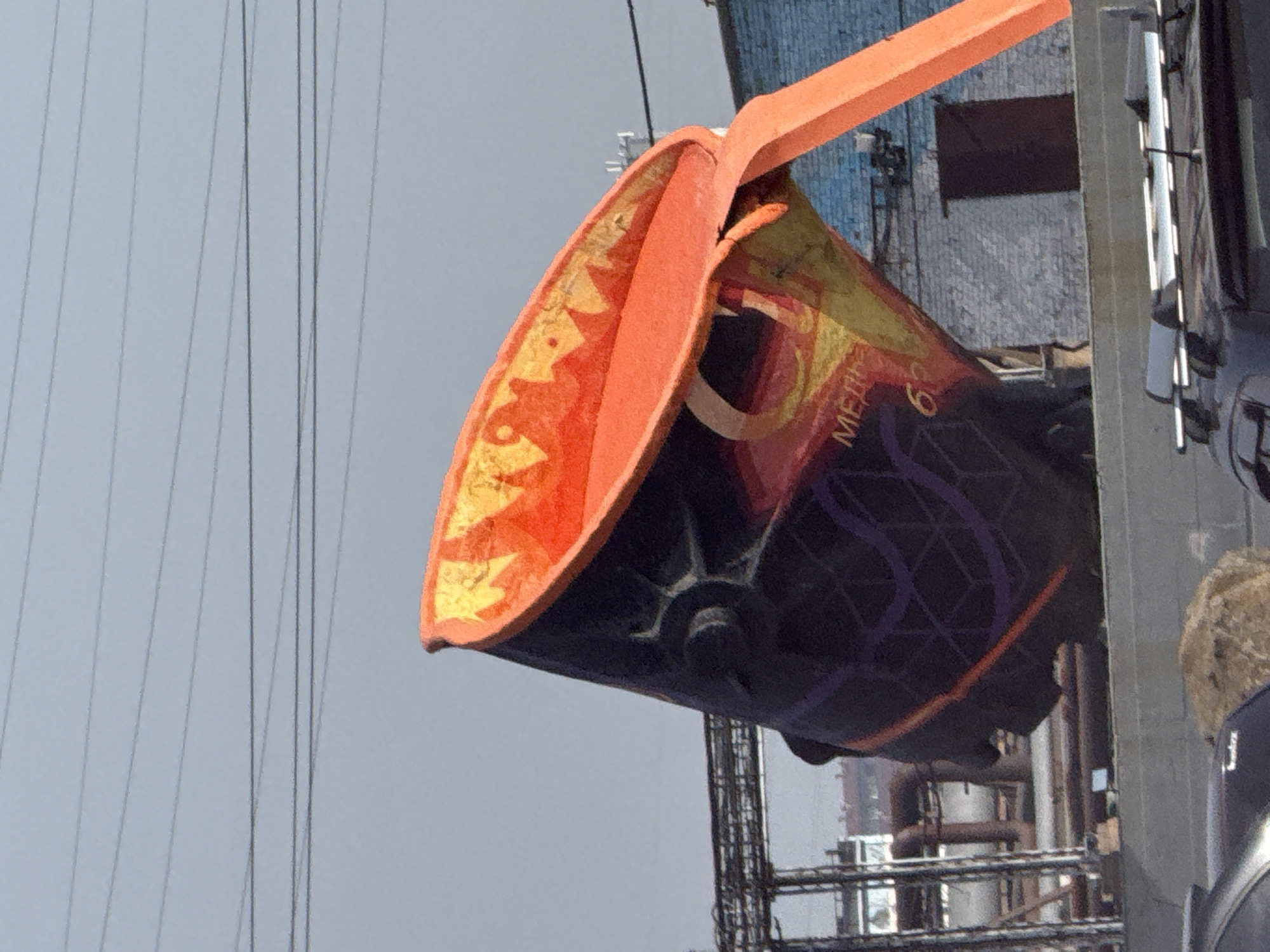 The image shows a close-up view of a large, tilted ladle, likely a sculpture, filled with what appears to be molten orange material. The ladle is decorated with dark purple and orange geometric patterns and some visible text, including "МЕДИ" and "63". It is positioned outdoors against a pale blue sky with several power lines running horizontally across the top. In the background, there are industrial structures with metal scaffolding and pipes, as well as a brick building with a dark door. The lower part of the image shows the roof of a dark-colored vehicle with roof racks. This appears to be a public art installation or monument located in the industrial city of Norilsk, Russia, a city known for its metallurgy and harsh climate.