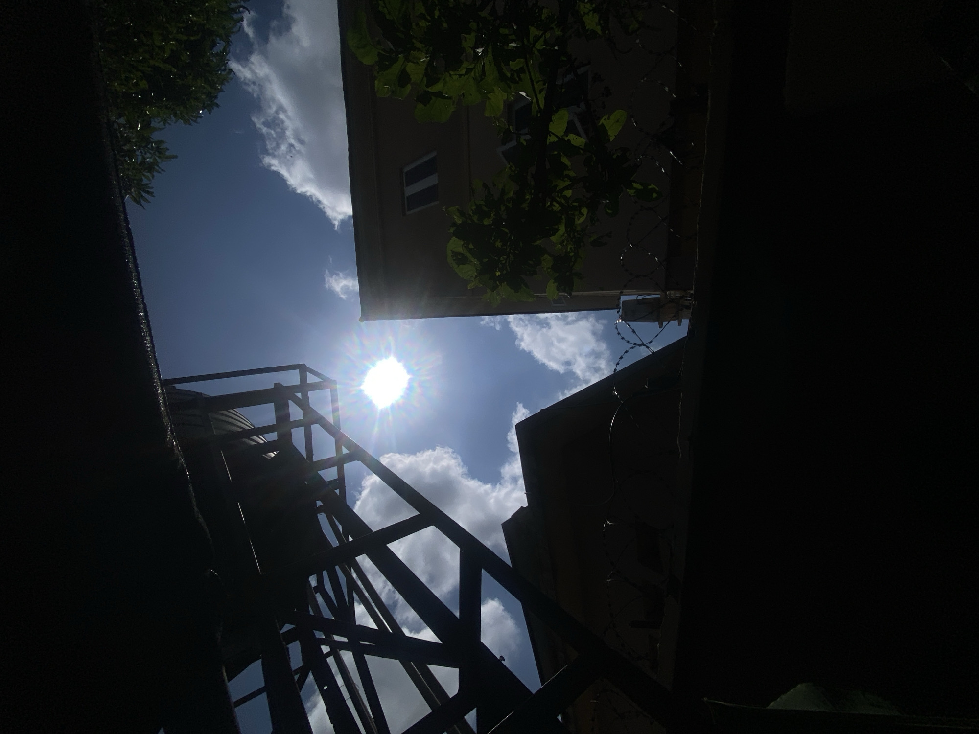 The image is a low-angle shot looking up towards the sky, featuring a bright sun with prominent lens flares. In the foreground, a dark, silhouetted structure, possibly a water tank tower, occupies the left side of the frame. To the right, a section of a building with a light-colored facade and a few windows is visible. Lush green foliage from a tree hangs over the building, partially obscuring it. Along the top of a dark wall in the lower part of the frame, there is barbed wire. The sky is a vivid blue, dotted with scattered white clouds. The overall impression is one of a sunny day in an urban or semi-urban environment, with a focus on the sky and architectural elements. The location is specified as Barden Yana, Nigeria.