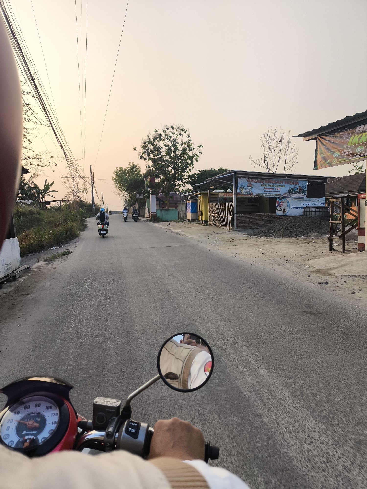 A first-person perspective video capture from a motorcycle shows a person riding on a paved road in Cilegon, Indonesia. The road is flanked by vegetation on the left and small buildings on the right. In the distance, several motorcycles are visible on the road. A sign with text in Indonesian is affixed to one of the buildings. The sky is overcast with warm hues, suggesting it is either sunrise or sunset. The rider's hand is on the handlebar, and a portion of the motorcycle's speedometer is visible in the lower left of the frame. The overall atmosphere is that of a casual ride through a semi-urban environment.