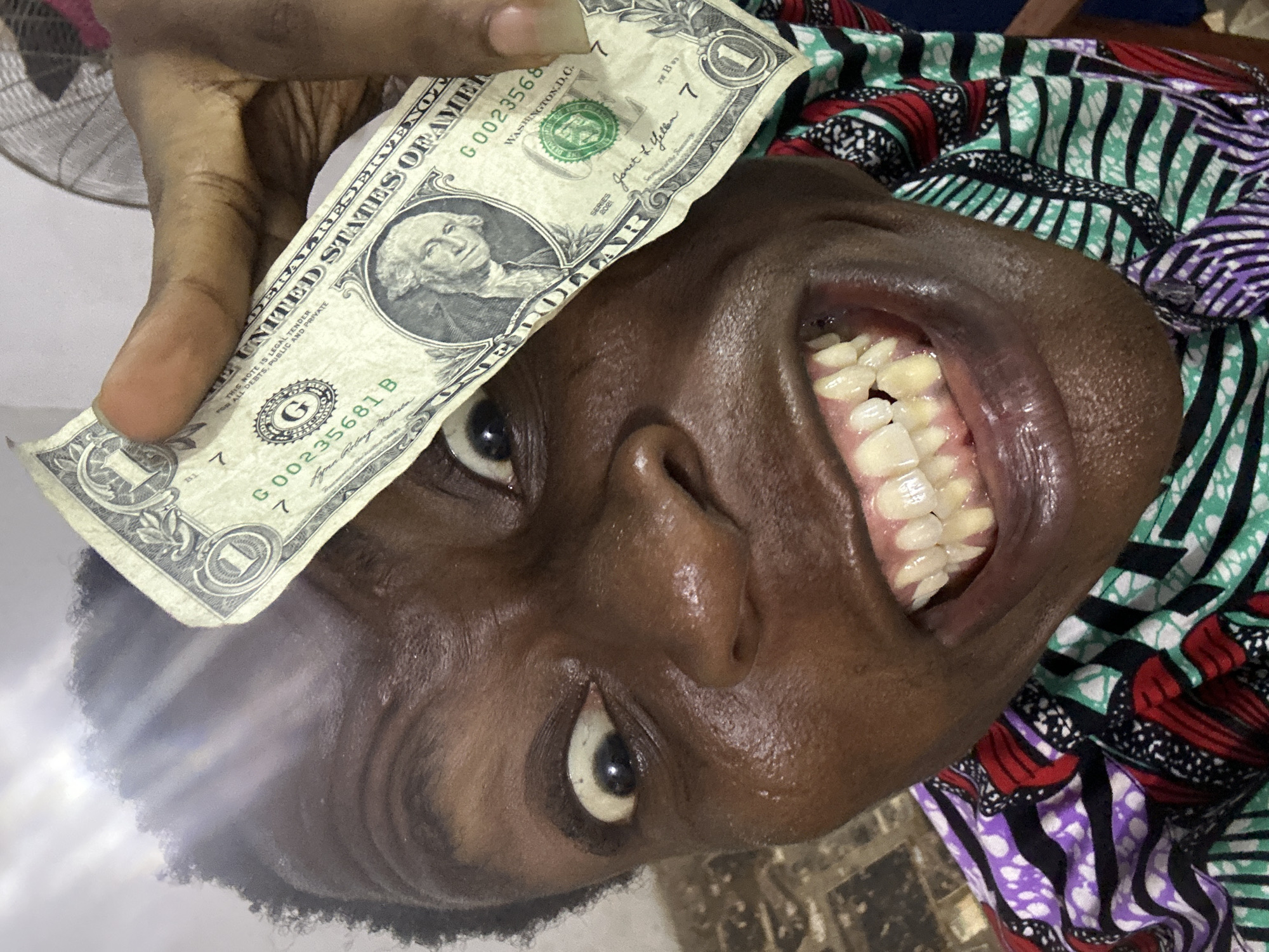 A close-up, rotated portrait shows a person with dark skin holding a creased US one-dollar bill near their face. The person's head is tilted, giving the impression of being upside down. One eye is visible, looking directly at the viewer, while the mouth is wide open, revealing numerous teeth. The teeth appear yellowish, with some visible gaps or dark spots between them, and the gums are dark. A bright lens flare is present in the lower left portion of the image.
The person's fingers grip the dollar bill over their forehead/hairline. Behind the person's head, colorful patterned fabric, possibly clothing, is visible with red, green, black, white, and purple elements. The background is blurry, suggesting an indoor setting, with faint hints of a white surface and possibly a fan.
Visible text on the one-dollar bill includes: "UNITED STATES OF AMERICA," "THIS NOTE IS LEGAL TENDER FOR ALL DEBTS, PUBLIC AND PRIVATE," the serial number "G00235681 B," "WASHINGTON D.C.," the Federal Reserve Seal with the letter "G," "SERIES 2021," and the signature of "Janet L. Yellen." The denomination "1" is visible in the corners of the bill.