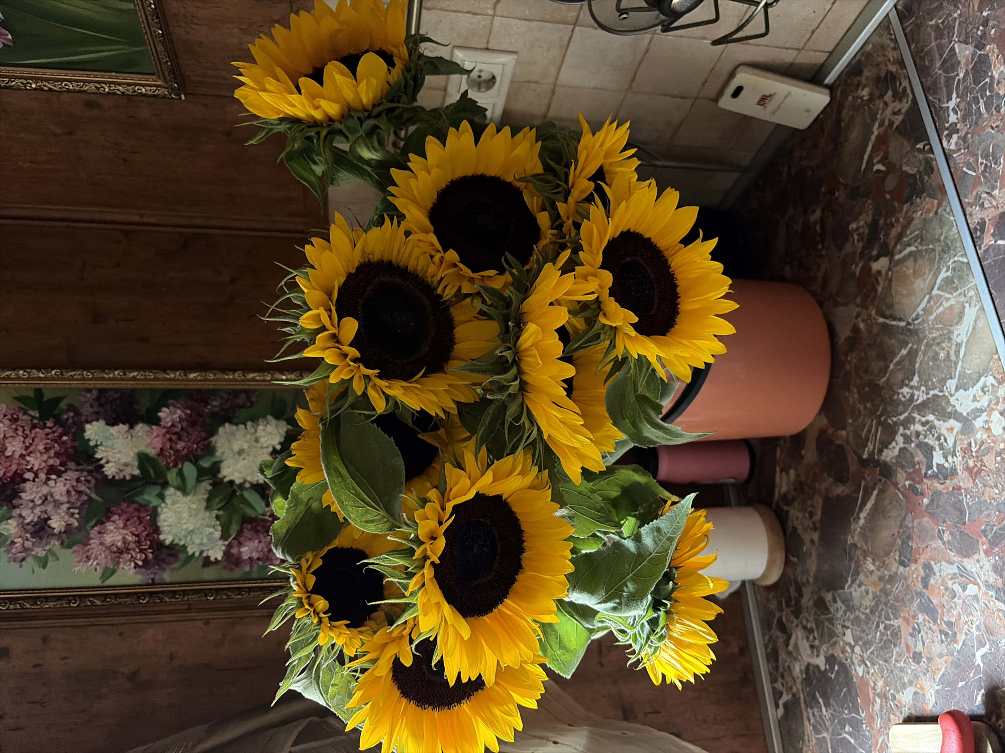 A bouquet of sunflowers is arranged in a large terracotta pot on a marble countertop. Behind the flowers, on the wall, is a framed painting of lilac flowers. To the right of the sunflowers, there is a light-colored electrical outlet. The scene appears to be an indoor setting, likely a kitchen, in Balashikha, Russia.