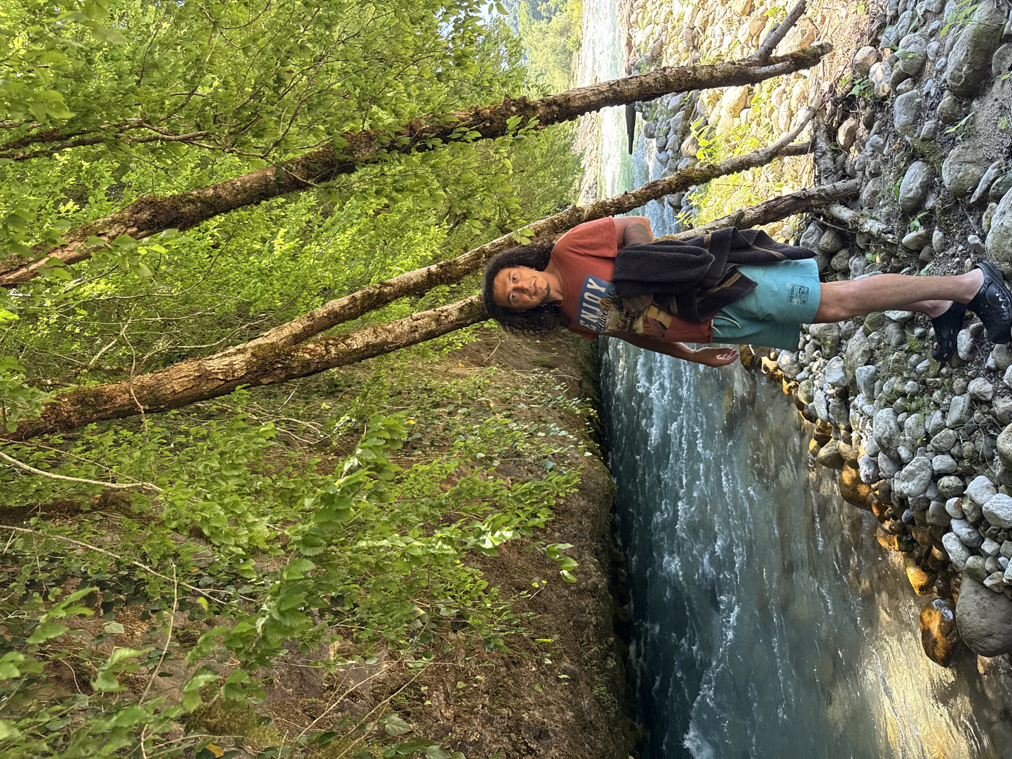 The media file depicts a person standing by a river in a natural setting. The person, with long, curly dark hair and a light complexion, is wearing a reddish-orange t-shirt with a blue graphic that reads "ENJOY Adventure" and light blue swim trunks. They are also wearing black slip-on shoes. A dark garment, possibly a jacket or towel, is draped over their shoulders.
The scene is a forest with lush green foliage and moss-covered trees. The river is on the right side of the frame, flowing with clear, turquoise water. The riverbank is lined with various-sized rocks and pebbles, some of which are illuminated by sunlight, suggesting it is daytime. The trees appear to be growing at an angle, with branches reaching out over the river and the bank.
The orientation of the image is portrait, with the river and the person appearing to be on a vertical plane. The person is looking towards the viewer with a relaxed expression. The overall impression is one of a peaceful, outdoor environment, possibly a recreational area for swimming or hiking. There are no other people visible in the frame. The location is identified as Abgara, Abkhazia. The lighting suggests it is a sunny day, and the abundance of green leaves indicates a warm season, likely spring or summer.