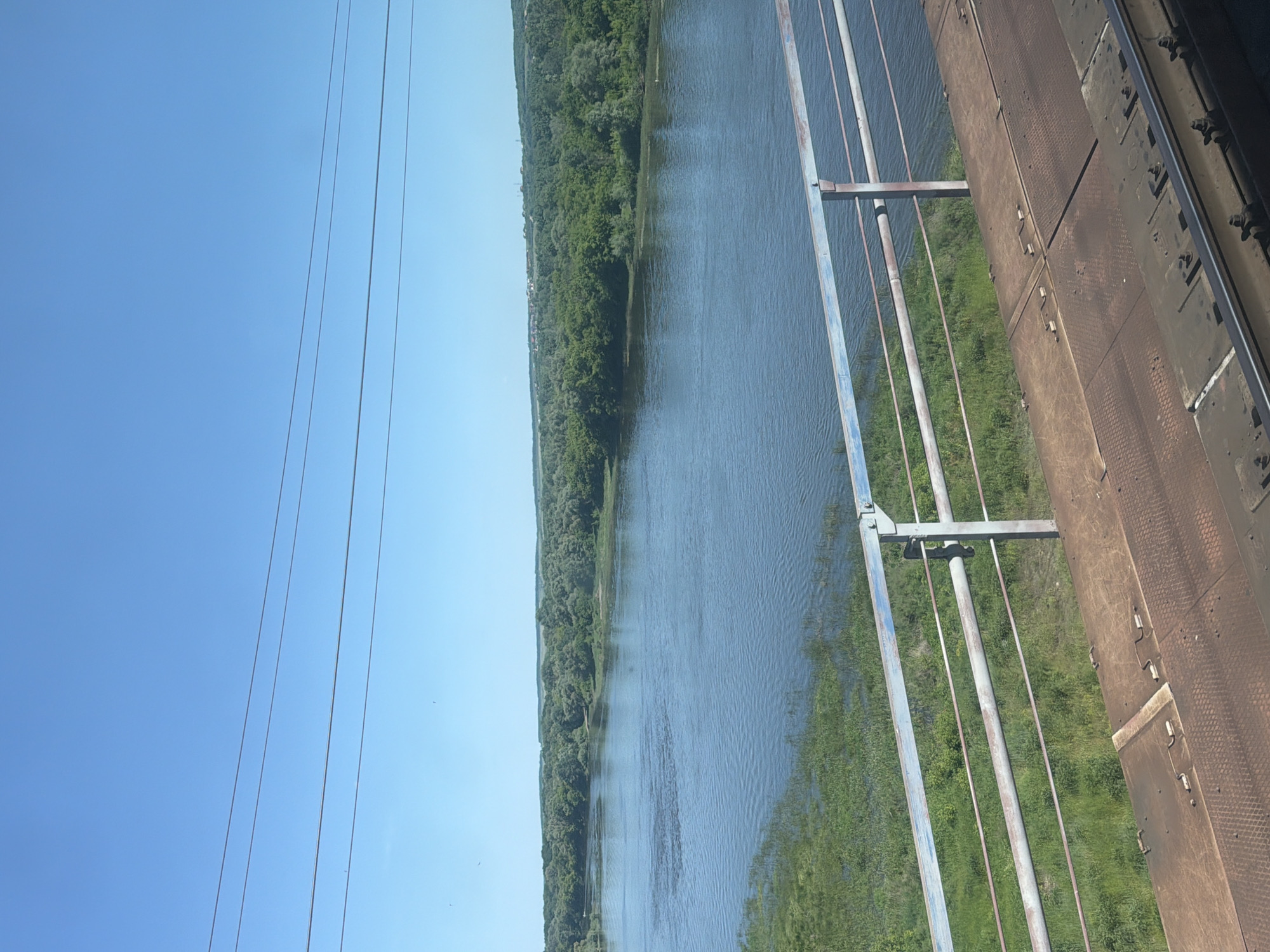 The image shows a wide, calm river with a lush green treeline along its banks. In the foreground, there is a bridge railing and train tracks, suggesting the photo was taken from a train. The sky is a clear, bright blue with a few thin power lines stretching across it. The scene appears to be a natural landscape with man-made infrastructure. There are no people or animals visible in the image. This scene is consistent with views from the railway bridge crossing the Don River in the city of Liski, Russia.