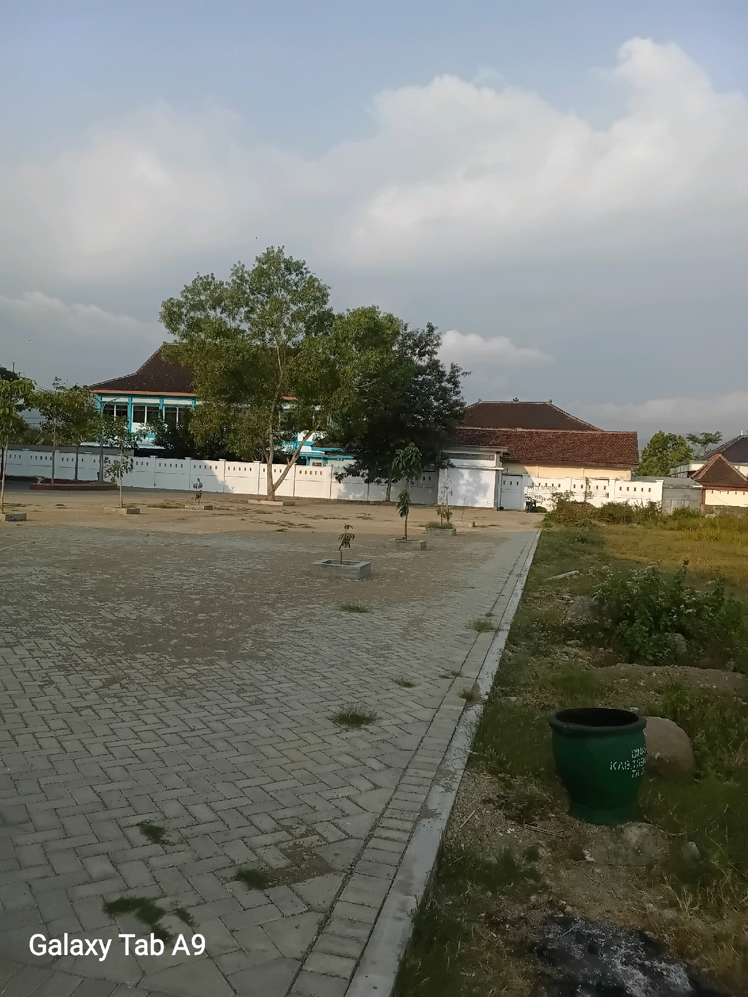 The image depicts an outdoor scene, possibly in the city of Trenggalek, Indonesia, with a focus on a paved area and a building in the background. The foreground is dominated by a light-colored, patterned paving stone surface, with some patches of grass and dirt visible. A sidewalk with a gray concrete border runs along the right side of the paved area. Beyond the paved area, a white wall with regularly spaced square openings separates the open space from the buildings.
Behind the wall, a two-story building with a blue facade and white trim is visible, featuring multiple windows. Another building with a terracotta tiled roof and a light-colored exterior is situated further to the right. Trees, some with full green foliage and others sparser, are scattered throughout the scene, with one notably large tree to the left of the blue building. The sky above is partly cloudy, with a mix of white clouds and blue sky, suggesting it is daytime. On the right side of the image, in the grassy area next to the sidewalk, a large green trash can with some text is placed. A rock is also visible next to the trash can. The overall impression is of a public space or institutional grounds, possibly a school or community center, in a tropical or subtropical climate.