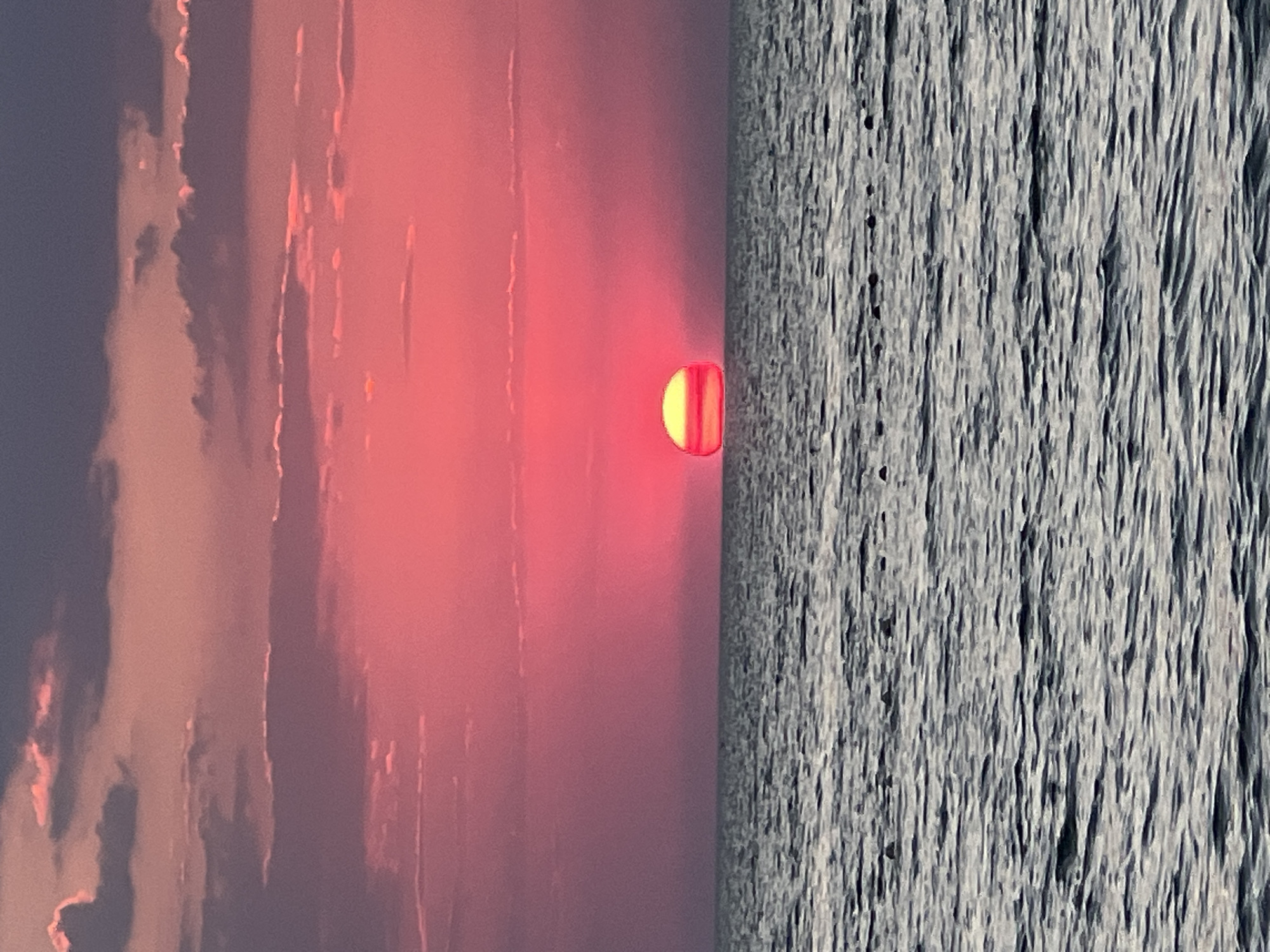 A sunset is captured over a body of water, likely the Black Sea, as viewed from the coast of Челядиновское сельское поселение, Ukraine. The sky is a vibrant gradient of pink and purple, with scattered clouds tinged with the same colors. A large, bright orange sun is setting on the horizon, appearing as a semi-circle just above the water. The water itself is a deep gray, with visible ripples and small, dark objects that could be buoys or small boats forming a line in the middle distance. The overall scene evokes a sense of calm and natural beauty.
