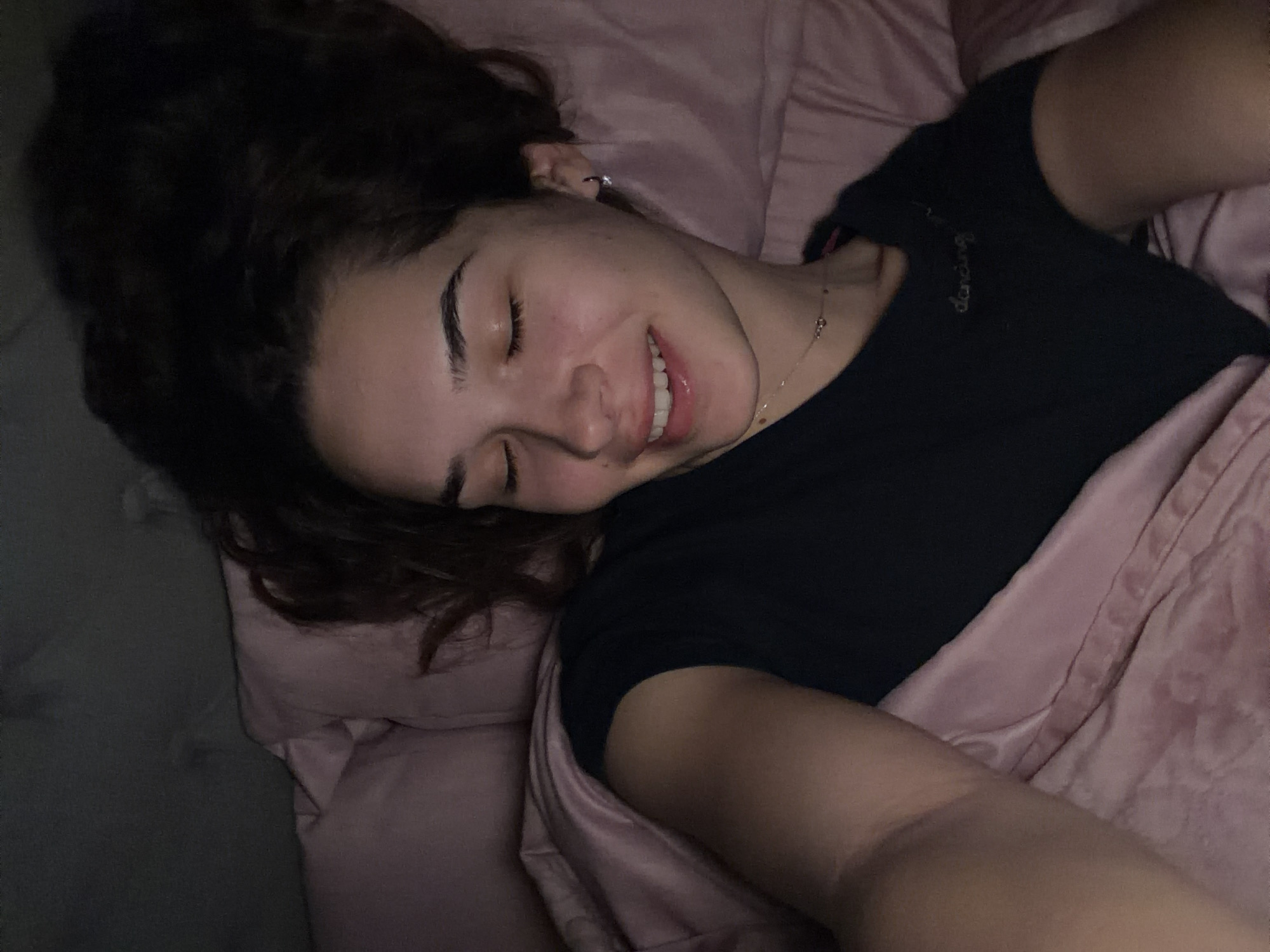 A woman is lying on a bed with pink bedding. She is wearing a black t-shirt with the word "dancing" on it. Her hair is dark and curly, and she is smiling with her eyes closed. She is wearing a necklace and earrings. The location is not discernible from the image, and Dobrich, Bulgaria, cannot be confirmed.
