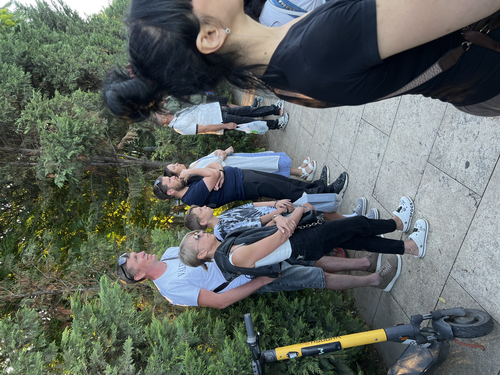 In Krasnodar, Russia, a group of people are captured in a vertical, eye-level shot, standing on a paved pathway lined with lush green cypress trees. The scene appears to be outdoors, likely in a park or public space, under the warm glow of what seems to be late afternoon or early evening light. The weather is pleasant, with dappled sunlight filtering through the trees, suggesting a clear or partly cloudy sky.
The group consists of at least six individuals, mostly adults, arranged in a line. They are all facing the same direction, with their bodies oriented horizontally in the frame due to the camera's orientation. The people are casually dressed, indicative of a relaxed outing. Visible clothing includes t-shirts, shorts, and jeans. One person wears white Crocs with cartoon characters.
The activity is not clearly defined, but their synchronized posture and gazes suggest they are looking at something or someone out of frame, perhaps participating in a group activity or posing for a photograph. There are no strong indicators of specific emotions, though the overall atmosphere seems calm and leisurely.
Notable details include a yellow electric scooter parked on the right side of the frame, its handlebars and wheel visible. The pathway is made of light-colored stone tiles. The cypress trees are dense and well-maintained, framing the scene. A faint inscription is visible on the yellow scooter, possibly a brand name or model number, though it's partially obscured. The image has a slight tilt, making the subjects appear to be lying down if viewed without considering the camera's rotation.