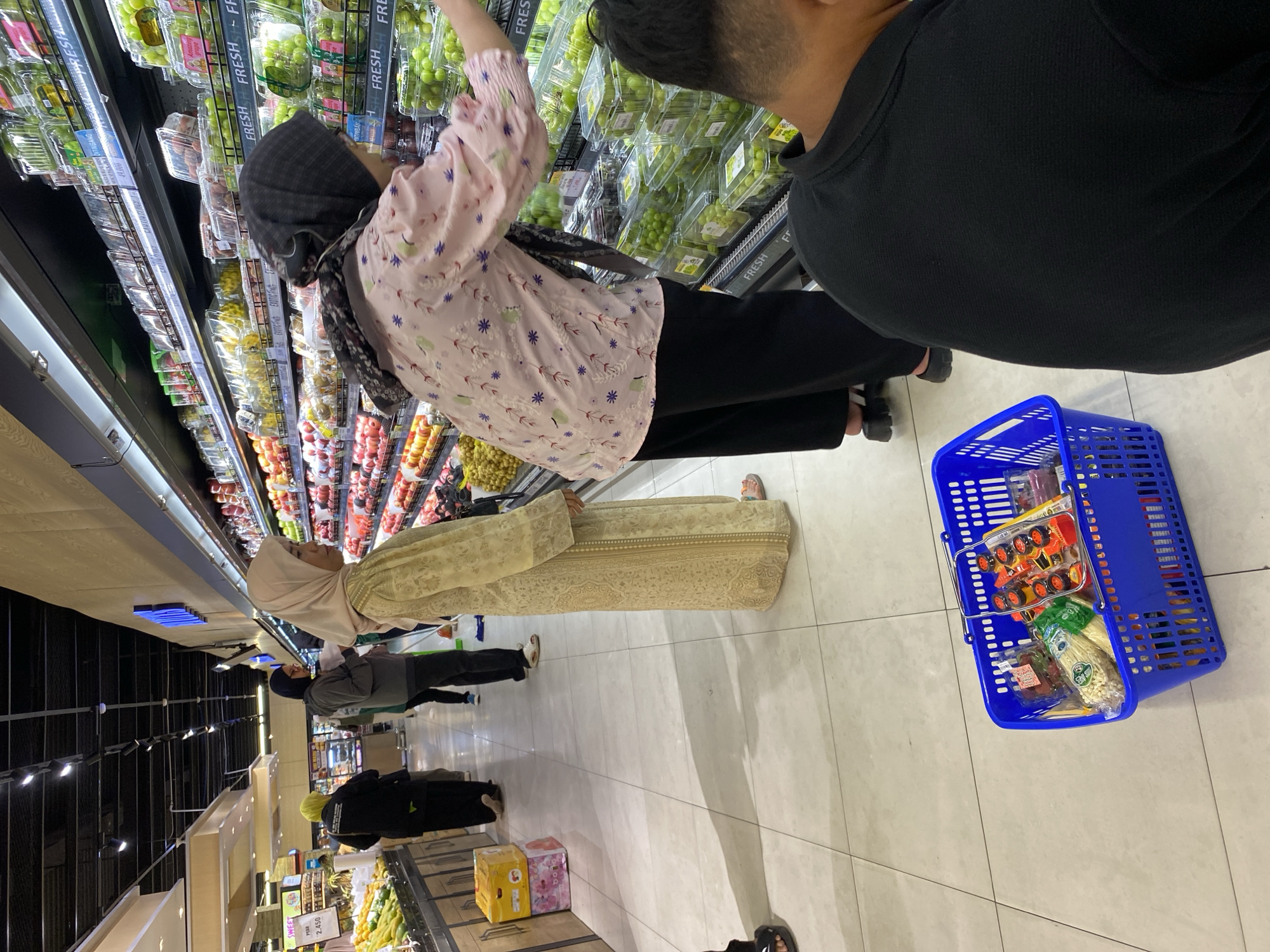 In the produce section of a brightly lit supermarket in Banda Aceh, Indonesia, a woman wearing a light-colored hijab and a patterned floor-length dress stands facing a display of fresh grapes. She is reaching out to select grapes from a clear plastic container. To her right, a person in a black t-shirt is partially visible from the back, also looking towards the produce. In the foreground, a bright blue shopping basket rests on the light-tiled floor, filled with groceries including packaged items such as strawberries, corn, and small toy-like objects. In the background, other shoppers are visible, one in a yellow hijab and black attire and another in a gray hoodie and black pants, browsing the shelves. The overall environment suggests a typical grocery shopping scene.
