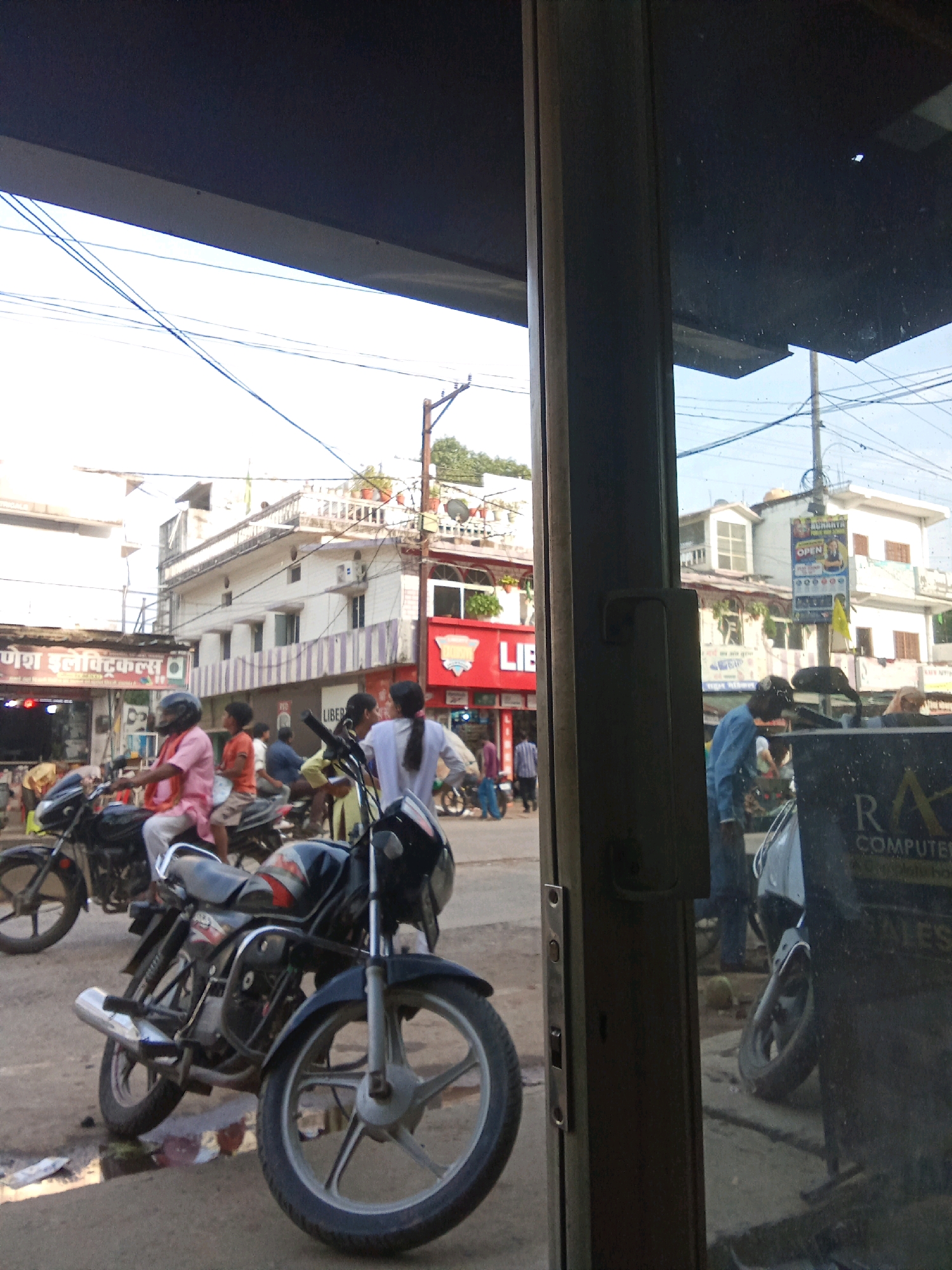 The image depicts a street scene in Sidhi, India, as viewed through a reflective glass surface. In the foreground, a black motorcycle with silver accents is prominently displayed. A person wearing a pink shirt and a black helmet is seated on a motorcycle in the middle ground, facing left. Other individuals are visible on motorcycles and walking along the street. Several buildings line the street, some with balconies and signs. Overhead, power lines crisscross the sky. The reflection in the glass shows a dark interior with some graffiti-like text, including the words "RA COMPUTER SALES". The overall impression is that of a busy urban environment.