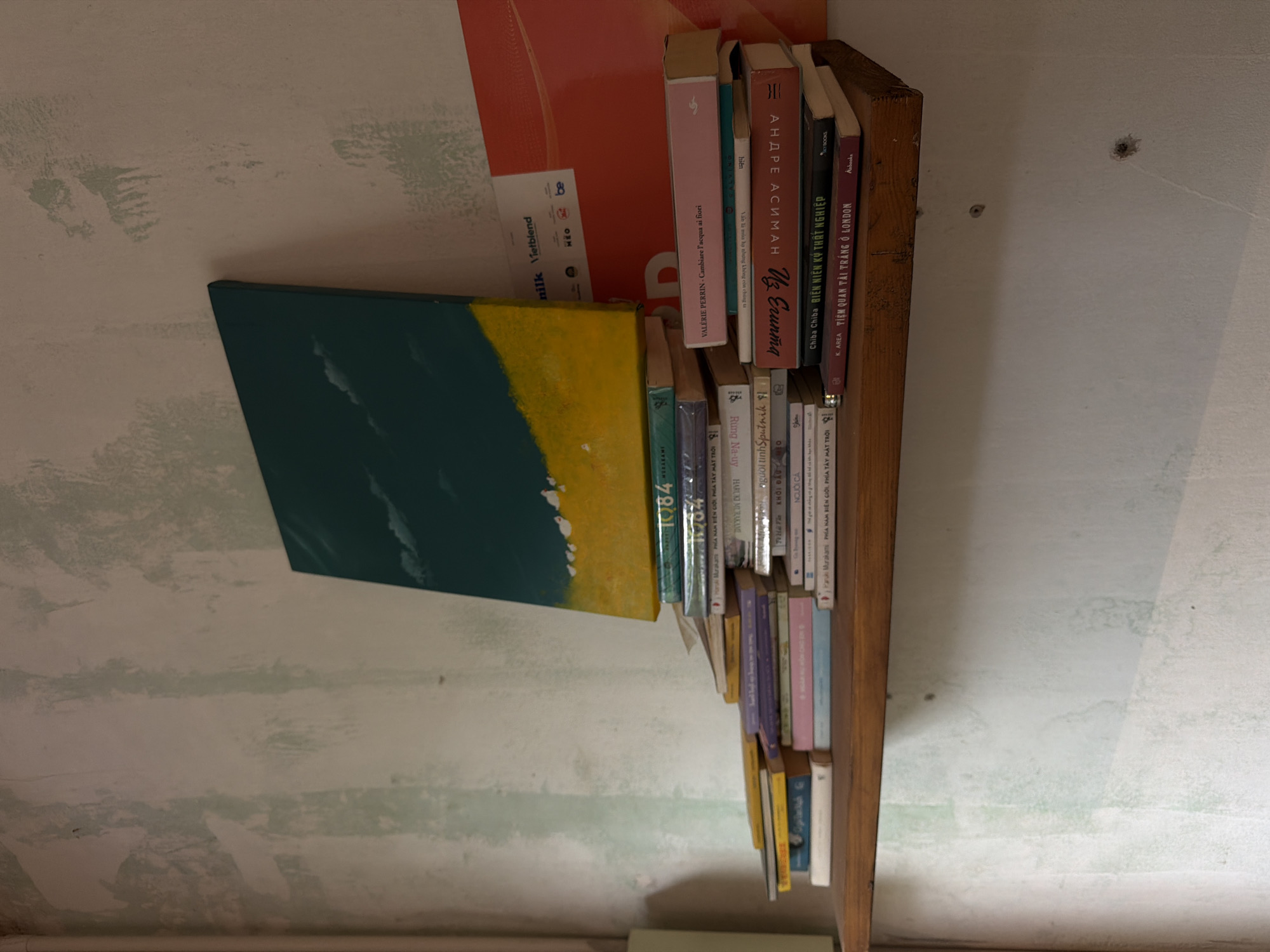 The image shows a wooden shelf mounted on a distressed, lightly painted wall. On the shelf, there is a stack of books and a small abstract painting. The painting, with a dark teal sky and a yellow ground with small white shapes, leans against the wall. The books are stacked in various orientations, some horizontally and some vertically, creating a dynamic arrangement. The wall exhibits signs of wear and tear, with patches of lighter paint and visible texture. The overall scene suggests a simple, possibly artistic, domestic setting. The location is identified as Đà Nẵng, Vietnam.