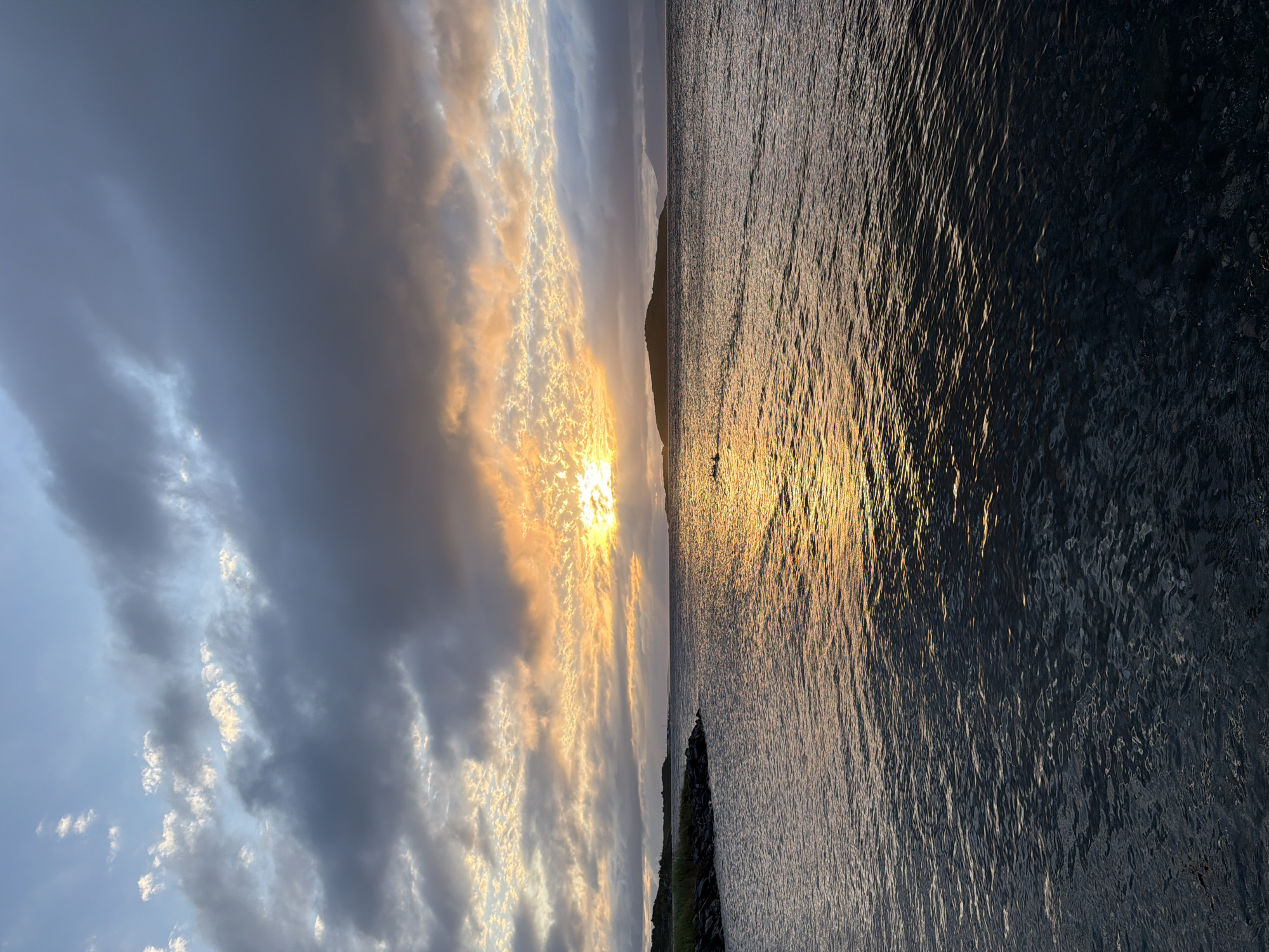 The image depicts a serene coastal scene at sunset in Harstad, Norway. The sky is filled with a dramatic display of clouds, with a portion of the sun breaking through, casting a warm, golden glow across the water. The sun's rays create a shimmering path on the ocean's surface, reflecting the vibrant hues of the sunset. On the left side of the frame, a rocky shoreline is visible, with some sparse vegetation. In the distance, a small island emerges from the water, adding depth to the landscape. The sea is calm, with gentle ripples disturbing its surface. The overall atmosphere is one of tranquility and natural beauty.
