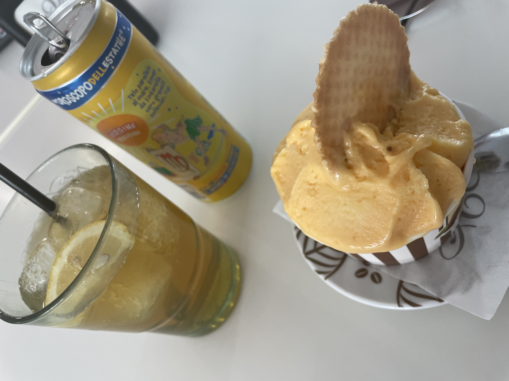 An overhead shot shows a glass of iced tea with lemon slices and a metal can of Estrella Damm beer next to a bowl of orange sorbet with a waffle cookie. The scene is set on a white table.