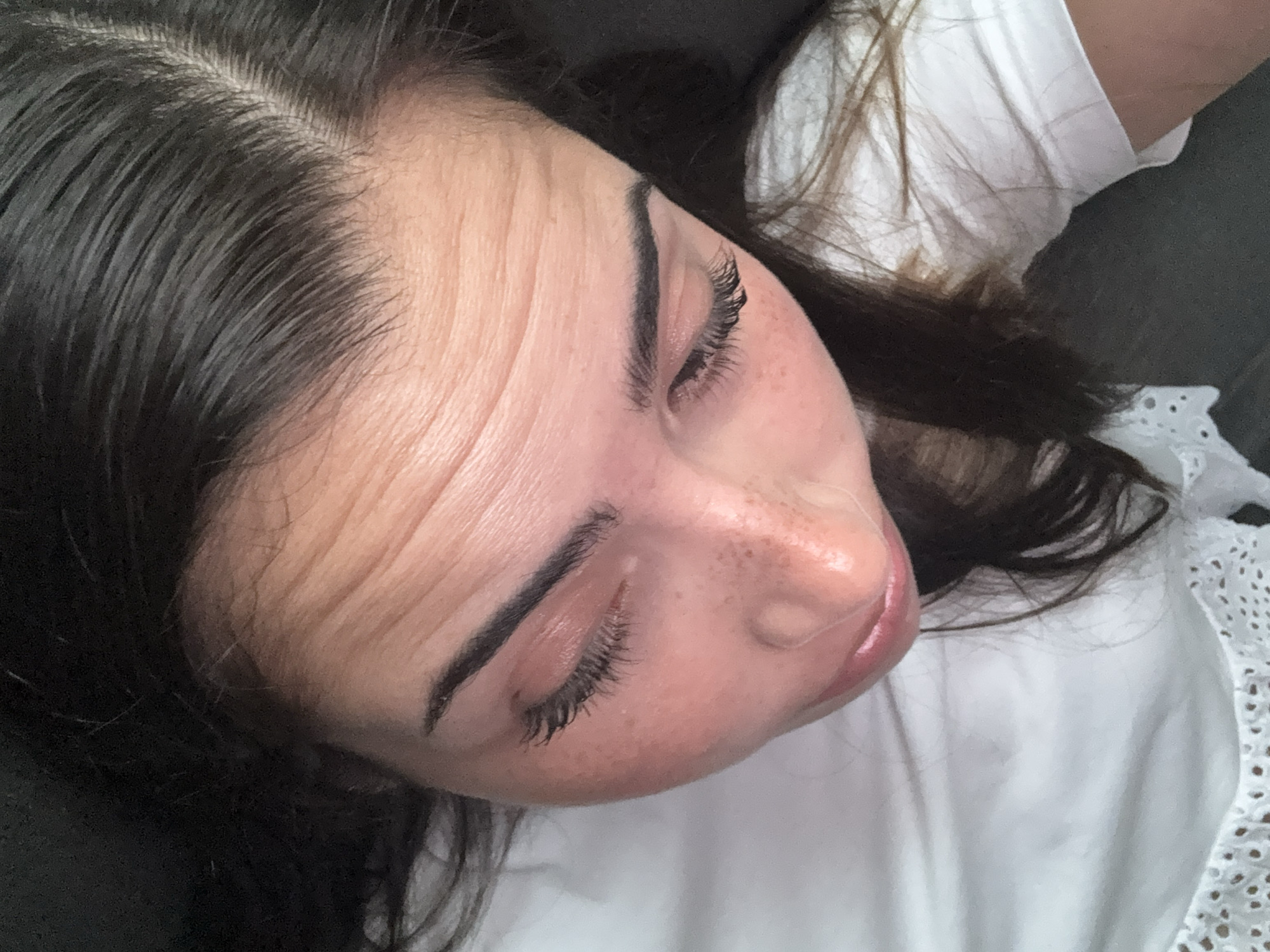 An overhead, close-up shot of a woman's face. The woman has dark brown hair, visible wrinkles on her forehead, and freckles on her face. Her eyes are closed, and she has dark eyebrows and eyelashes. She is wearing a white shirt. The background is a mix of dark and light colors. There is no information available to identify the location as San Bernardino Tlaxcalancingo, Mexico.