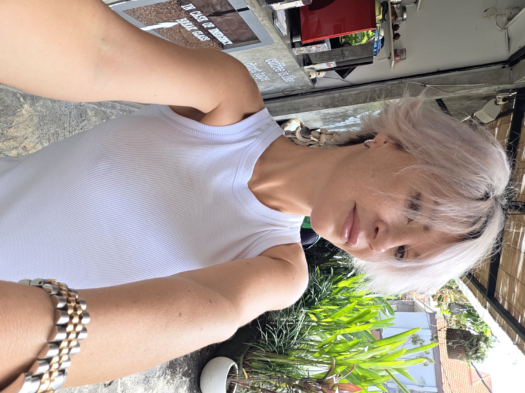 In North Kuta, Indonesia, a woman with short, light-colored hair poses for a selfie. She is wearing a white ribbed tank top and a two-tone watch on her left wrist. The background suggests an outdoor cafe or shop. To her left, a red espresso machine is visible, along with shelves stocked with jars. A sign on the wall behind her reads "coffee is like duct tape," and another partially visible sign says "IN CASE OF EMERGENCY BREAK GLASS." Lush green plants in a white pot are to her right, and parts of buildings with orange roofs are visible in the distance. The overall atmosphere is casual and sunny.