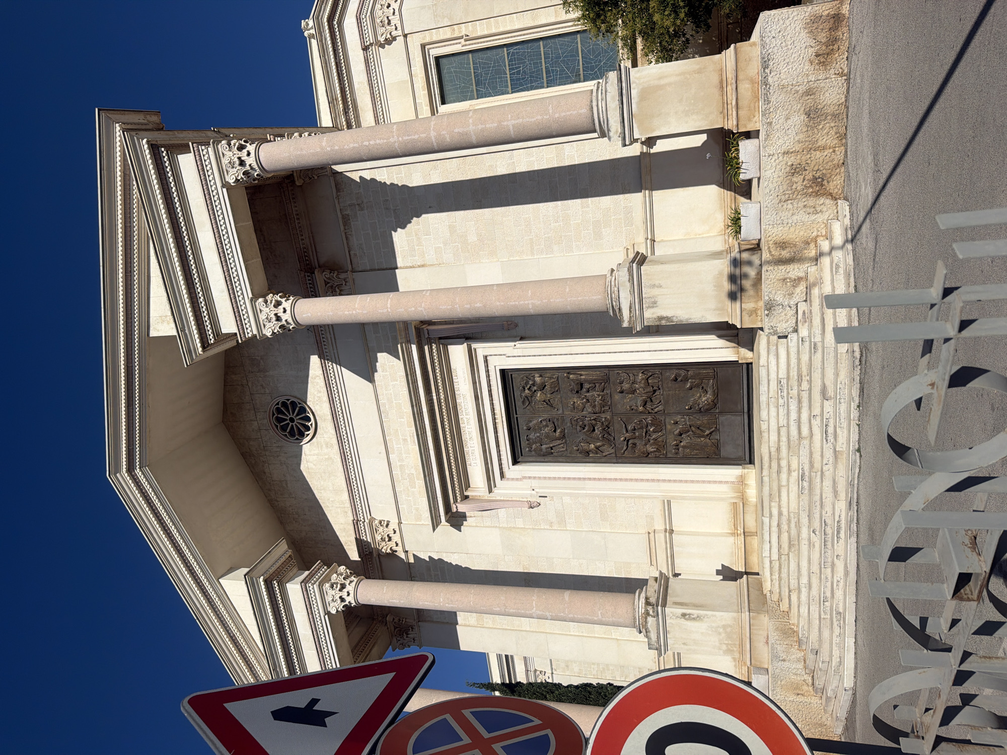 The image shows the facade of a building with neoclassical architecture, likely a church or public building, under a clear blue sky. The building is constructed of light-colored stone blocks and features a pedimented portico supported by Corinthian columns. A large, ornate bronze door is visible in the center, flanked by the columns. Above the door, an inscription is present. To the left of the entrance, a set of stone steps leads up to the building. In the foreground, on the left, a series of traffic signs are visible, including a directional sign with an arrow and two circular signs, one with a blue circle and red cross, and another with a red circle and a black number "70" with a diagonal line, indicating a speed limit. A metal fence with a decorative, curved design is in the lower foreground. The ground is paved with asphalt. A slender tree is visible to the right of the building, partially obscuring a stained-glass window. Two rectangular planters with small plants are placed on a stone ledge to the right of the steps. The lighting suggests it is daytime, with strong shadows cast by the architectural elements and the fence. The building's design and the overall atmosphere evoke a sense of historical and cultural significance. While the specific location in Bari, Italy, cannot be definitively determined solely from the visual elements in this image, the architectural style is consistent with structures found in Italian cities.