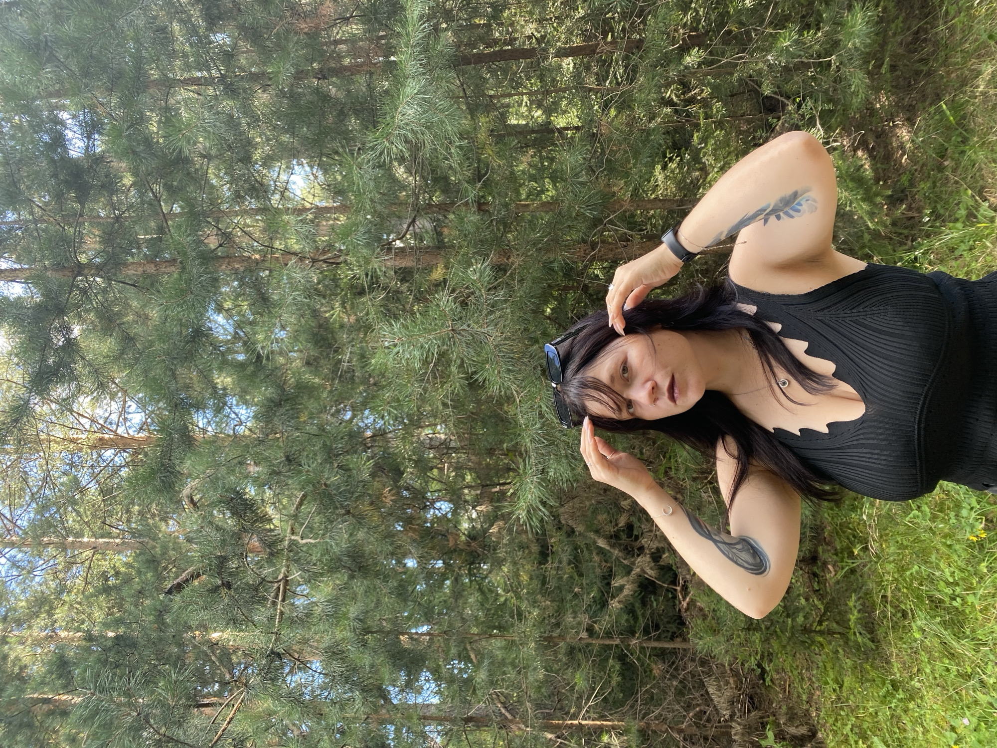 A young woman with dark hair and bangs, wearing a black tank top and sunglasses perched on her head, stands in a forest. She has tattoos on her arms, a silver necklace, and a black smartwatch. Her hands are raised to her head, and she is looking directly at the camera. The background is filled with tall pine trees and lush green foliage. The woman is positioned in the center of the frame, with the forest occupying the majority of the image. The scene appears to be a sunny day, with natural light filtering through the trees. The image was taken in the city of Fokino, Russia.