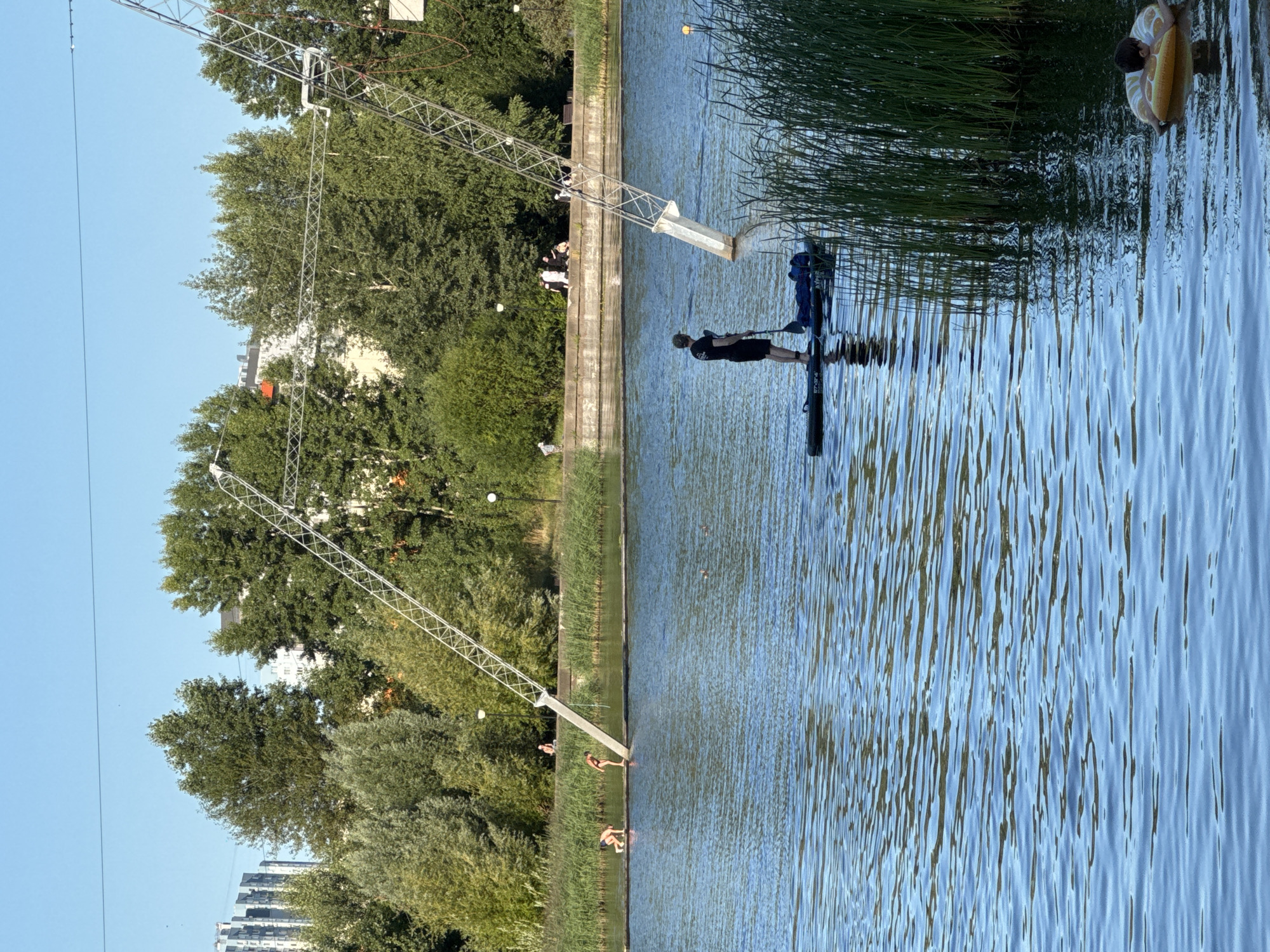 The image depicts a sunny day at a recreational area, possibly a wake park or a lake with water sports facilities, located in Nizhny Novgorod, Russia. In the foreground, a person is paddleboarding on a calm body of water. The paddleboarder, dressed in black shorts and a dark t-shirt, stands with a paddle in hand, facing away from the camera. Behind them, tall reeds line the edge of the water. To the right, a young child floats on a yellow inflatable ring. In the mid-ground, a wooden platform extends into the water, with several individuals near it. Two people, one in a black swimsuit and another in a red bikini, are positioned on the platform, appearing to be preparing to enter the water or observing others. Further back, on the shore, a group of people are gathered, and more individuals are visible further along the embankment. Towering metal structures, likely part of a wakeboarding cable system, rise prominently on the right side of the image, their angled frames reaching towards the clear blue sky. The background features a line of lush green trees and buildings, including apartment blocks, suggesting an urban or suburban setting within Nizhny Novgorod. The water surface reflects the bright sky and surrounding environment, creating a dynamic texture.