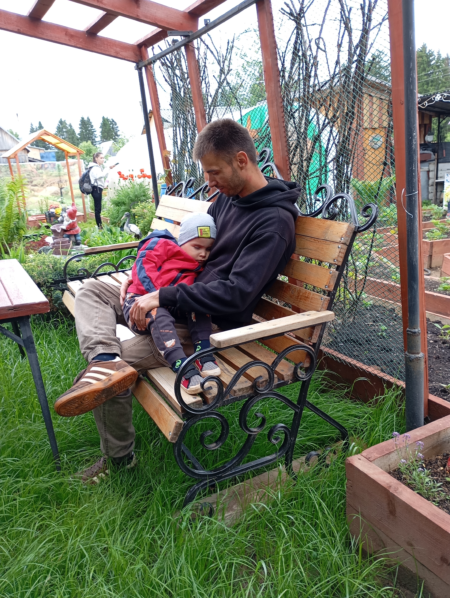 The image depicts an outdoor garden scene in the city of Фоки, Russia, featuring a man and a child on a bench under a wooden pergola on an overcast day.
In the foreground, a man with short, dark hair and stubble is seated on a wooden bench with black wrought iron embellishments. He wears a black hooded sweatshirt and olive-brown cargo pants. His left foot, clad in a brown Adidas sneaker with white stripes and a gum sole, rests on his right knee. A young child, appearing to be a toddler or preschooler, is asleep on the man's lap, facing his chest. The child wears a red hooded jacket, dark patterned trousers, a light grey beanie with a yellow patch, and black and white checkered slip-on shoes with red accents. The man's gaze is directed downwards towards the child.
To the left of the bench, a simple wooden table with dark metal legs is visible. The ground around the bench and table is covered in lush, somewhat overgrown green grass.
In the middle ground, further back in the garden, a woman with light-colored hair is standing. She wears a light-colored long-sleeved top, dark pants, and a dark backpack. She holds a dark object, possibly a smartphone, in her right hand and appears to be looking towards the left. This area of the garden features various plants, including ferns and red poppies, and garden ornaments such as red-hatted gnomes and a grey bird statue.
To the right of the bench, there are raised wooden garden beds containing dark soil and young green plants, with a few small purple flowers visible in one bed. Behind the bench and garden beds, a chain-link fence is present, partially obscured by bare tree branches. In the distant background, several residential structures are visible, suggesting a village or suburban environment.