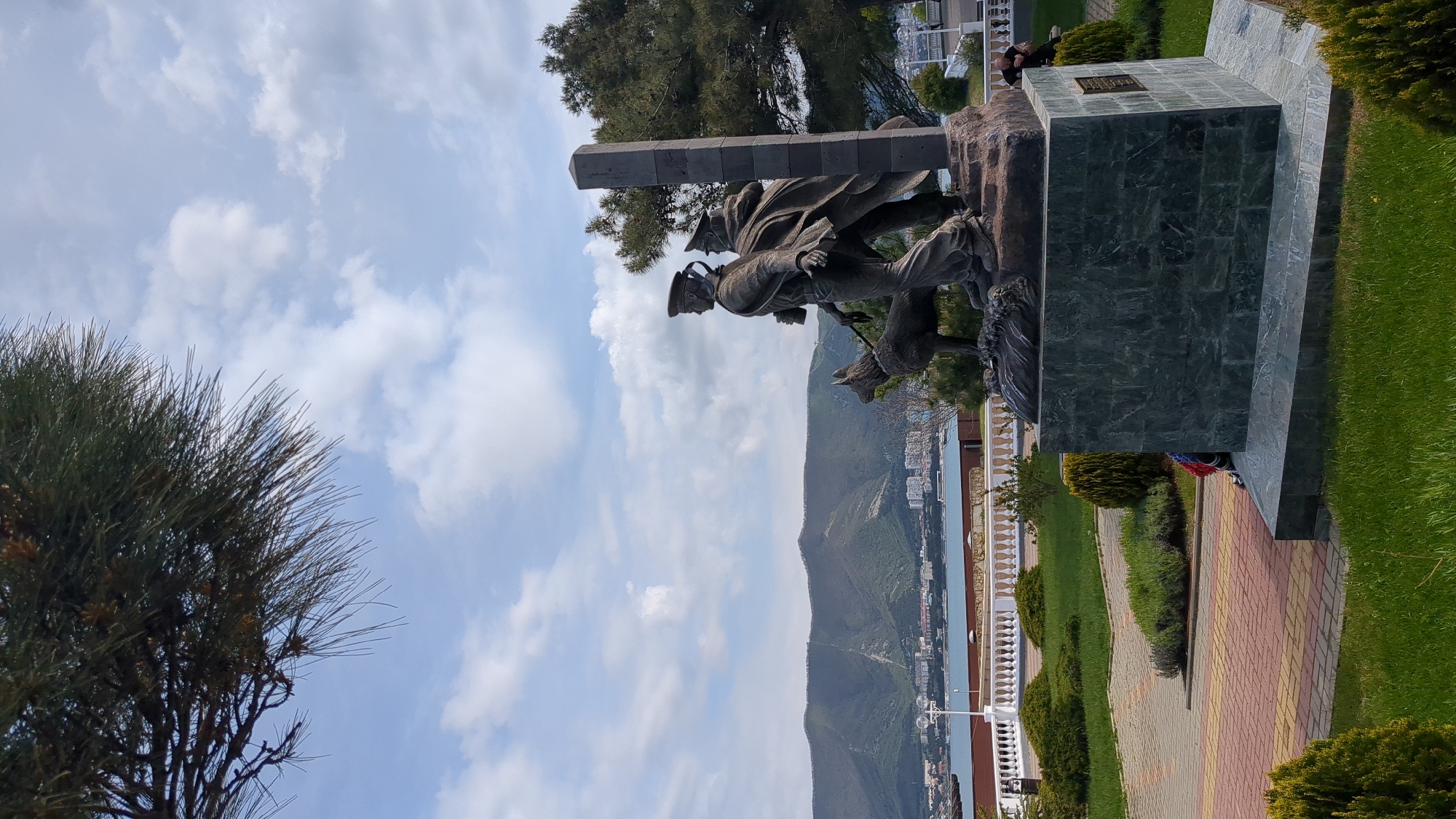 The image displays a prominent monument overlooking a picturesque coastal landscape. In the foreground, a bronze-colored sculpture features two male figures and a dog. The central figure, dressed in what appears to be a military uniform with a cap, stands tall, holding a rifle-like object. A second figure is partially visible behind him, and a dog walks beside the lead figure.
The sculpture rests upon a substantial base constructed from dark green, veined marble or stone. An inscription plaque is affixed to the side of this base, and a wreath adorned with red, white, and blue ribbons lies at its foot. A person is visible in the distance, sitting near the monument.
The monument is set within a meticulously maintained park. Lush green grass covers the ground, interspersed with manicured bushes and a pathway paved with colorful interlocking bricks in shades of red and yellow. Tall, needle-leaved trees are visible on the left and right, framing the upper portions of the scene against the sky.
The background unfolds into an expansive vista. A wide body of water, likely a bay or sea, stretches across the mid-ground, with a white balustrade separating the park from the view. Along the distant shoreline, a city with numerous buildings is nestled at the base of large, verdant mountains that rise dramatically into the horizon.
The sky above is bright blue, dotted with scattered white cumulus clouds, indicating clear and sunny daytime weather. The overall impression is one of a solemn memorial site situated in a picturesque, well-maintained coastal environment, consistent with Gelendzhik, Russia.