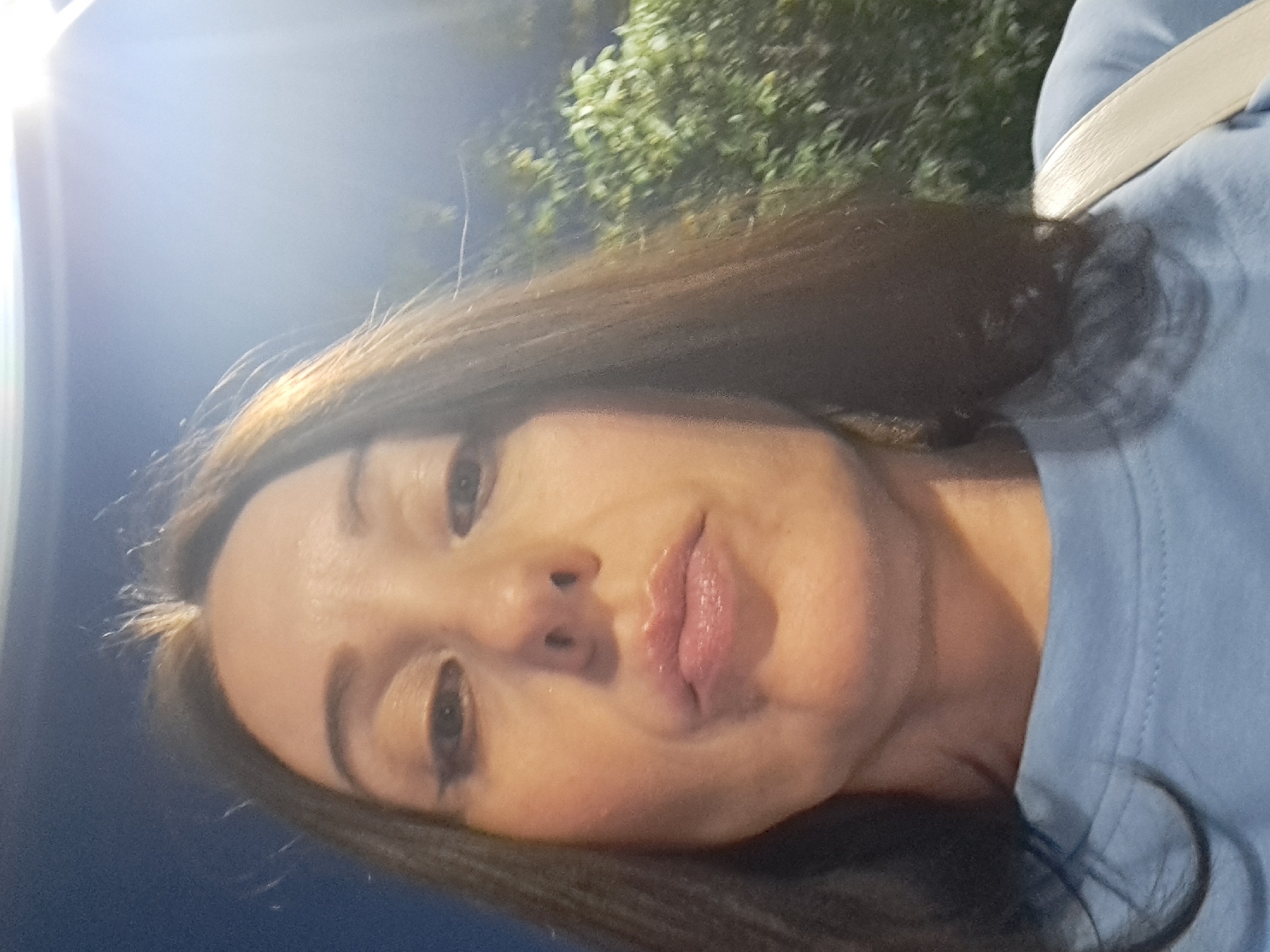 A low-angle, selfie-style photograph shows a woman's face and upper torso, taken outdoors at night. She has long, dark brown hair and is wearing a light blue t-shirt and a light-colored strap across her shoulder, likely from a bag. Her makeup includes winged eyeliner and lip gloss. The background is dark, with some blurred greenery visible on the right and a bright light source in the upper right corner, suggesting a streetlamp or artificial lighting. The scene is not identifiable as being in Irkutsk, Russia.