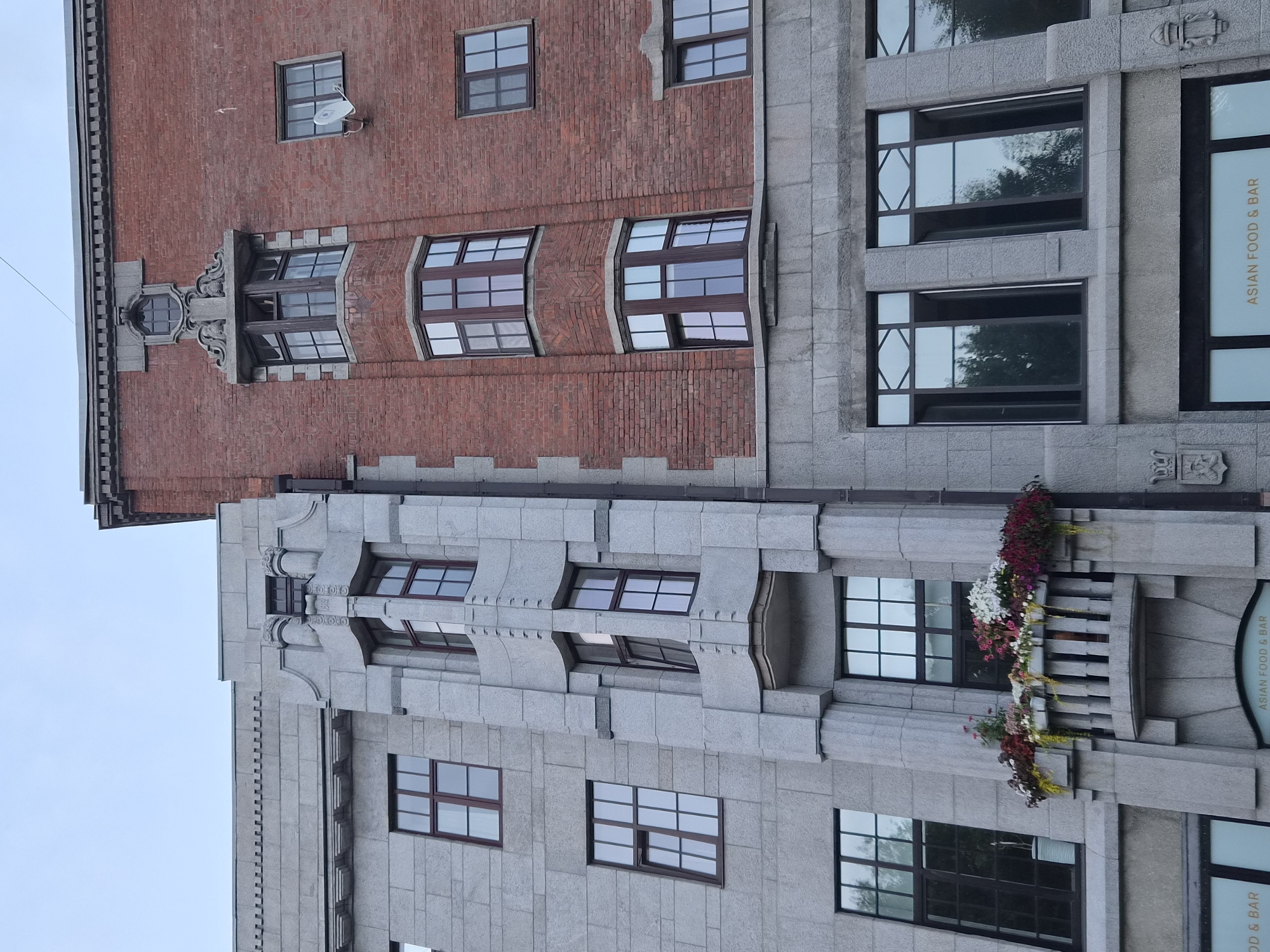 This is a low-angle, outdoor, full-shot photograph of the facade of two adjacent buildings, likely in Vyborg, Russia. The building on the left is constructed with light grey granite blocks, featuring a prominent vertical element with decorative vertical fluting and a small balcony adorned with potted flowers. The building on the right is constructed of red brick and has a more ornate architectural style, with elaborate stonework around some of the windows, including a bay window on the upper floors and a smaller circular window at the very top. Several windows are visible across both buildings, most of them multipane. The sky is overcast and appears light blue. There are no people or animals visible in the image. The scene appears to be a street view of urban architecture.