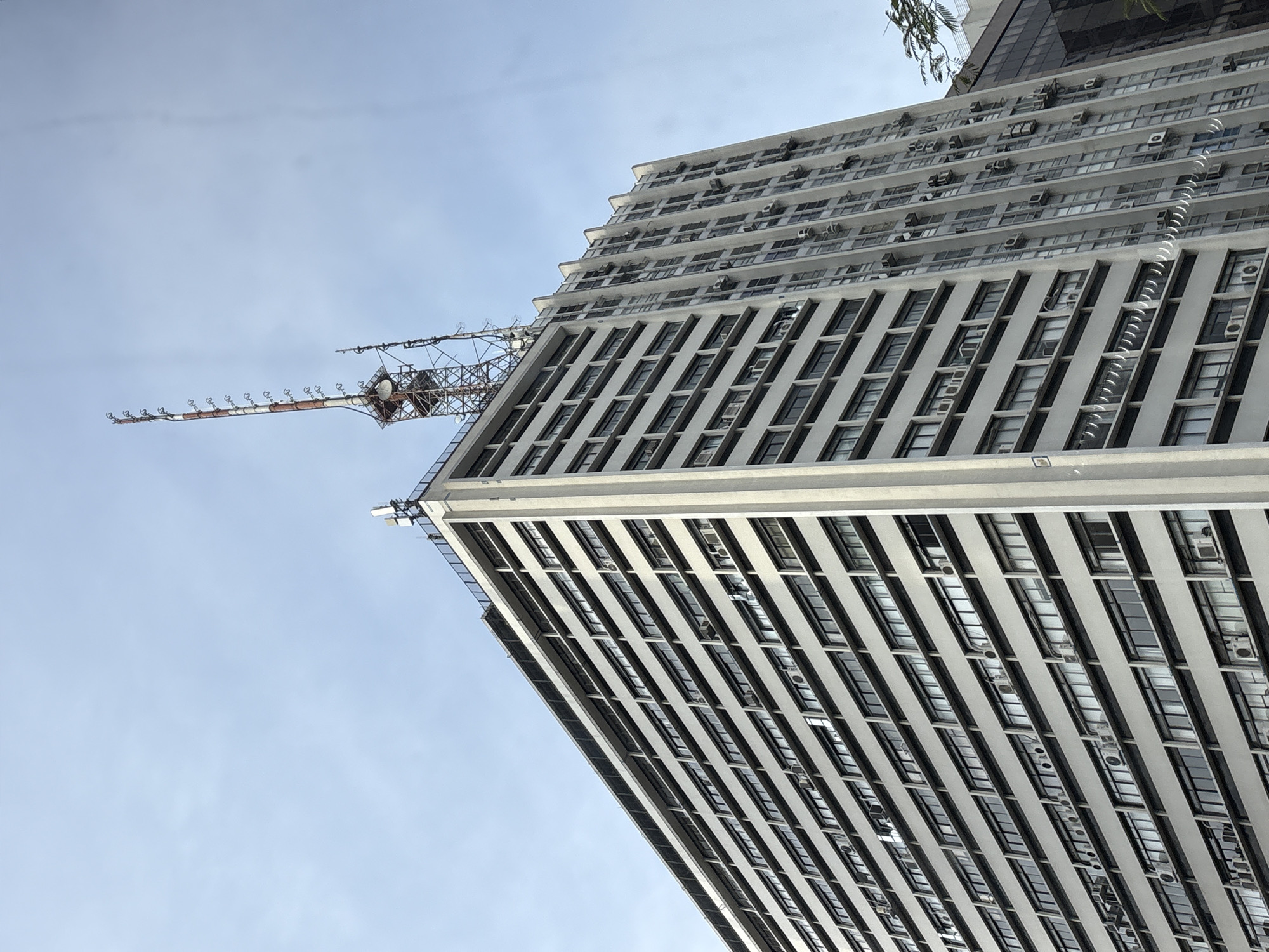 The image shows a low-angle view of the Edifício Martinelli, a historic skyscraper located in the center of São Paulo, Brazil. The building is tall and rectangular with a facade characterized by numerous windows and rows of air conditioning units. At the top of the building, a large radio tower with multiple antennas is visible against a clear, pale blue sky. The tower is a prominent feature, suggesting the building serves telecommunications purposes. The building's architecture is utilitarian, with repeating geometric patterns of windows and the concrete structure. The surrounding environment is not clearly visible, but the presence of the skyscraper indicates an urban setting. No people are visible in the image.