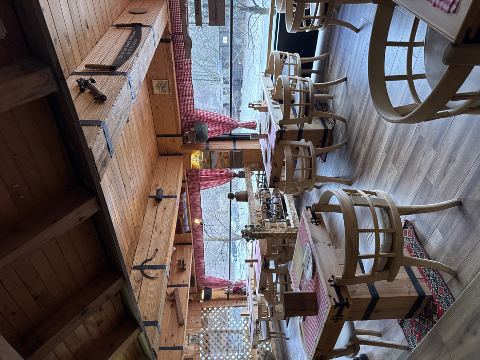 The image captures the interior of Restoran Amaret in Jabuka, Montenegro, depicting an unoccupied dining area. The room is constructed primarily of wood, featuring a sloped ceiling with exposed wooden beams, some reinforced with dark metal strapping and adorned with various antique-style tools or implements. The walls are paneled in wood. Large rectangular windows, framed by red and white checkered curtains tied back, line the far wall, providing a view of an overcast outdoor scene with bare trees and distant vehicles. The floor is tiled with a greyish-brown wood-plank pattern.
The dining area contains multiple sturdy, rectangular wooden tables. Several tables are set with red and white checkered placemats, white plates, cutlery (fork and knife), and glasses. Small wooden menu stands, one clearly displaying "AMARET," are present on various tables, and the name "AMARET" is carved into the side of at least one table. Numerous light-colored wooden chairs with round backs and cushioned seats are arranged around the tables. A decorative wooden structure incorporating an ornate metal element, possibly a wine rack, is positioned near the center-right windows. A pendant light fixture hangs from a ceiling beam in the mid-left area. No individuals are present in the scene, and no actions are depicted.