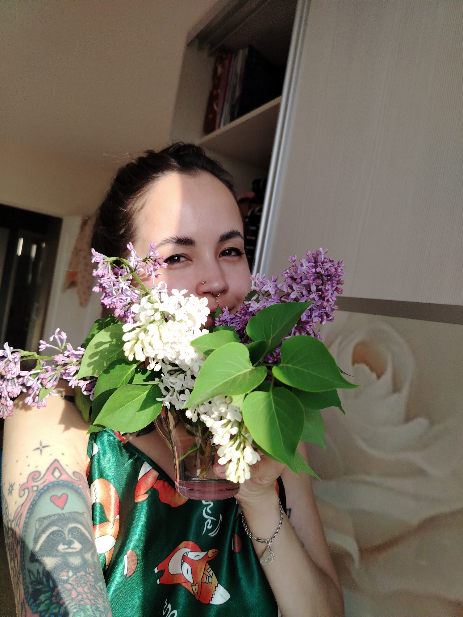 A woman is holding a vase filled with lilac flowers. She has dark hair pulled back and is wearing a green satin top with a pattern of orange and white foxes. She has a nose ring and a tattoo on her arm of a raccoon inside a frame. The background includes a white closet with books on a shelf. The location is inside a building, but the city is unknown.