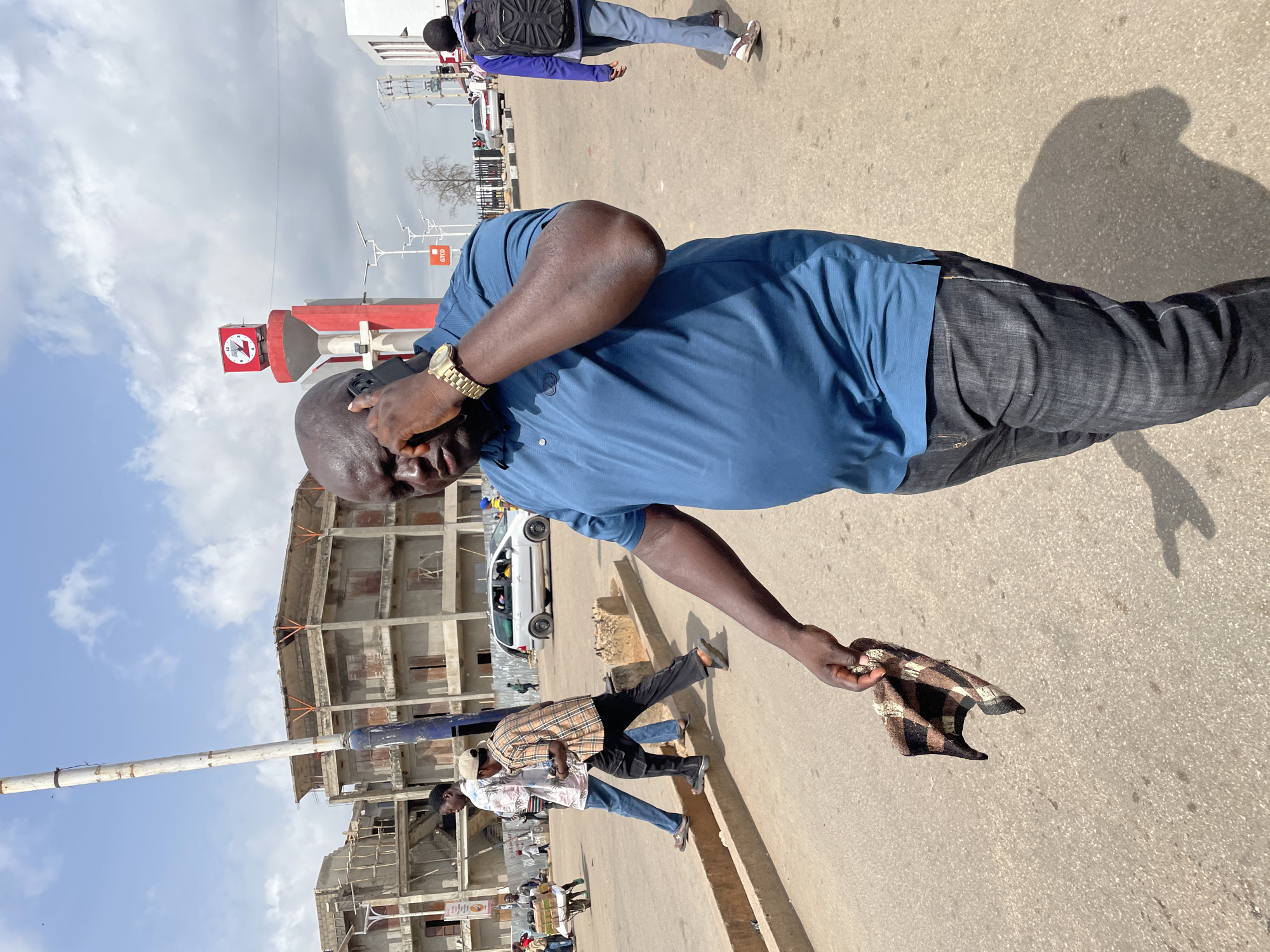 A man in a blue shirt and dark pants is standing on a paved street, holding a patterned cap in his right hand and a black object to his ear with his left hand. He is wearing a gold watch. Behind him are pedestrians and a white car. There's an unfinished building under construction in the background. A red and white sign is visible in the distance. The location appears to be in a city, likely Jos, Nigeria.