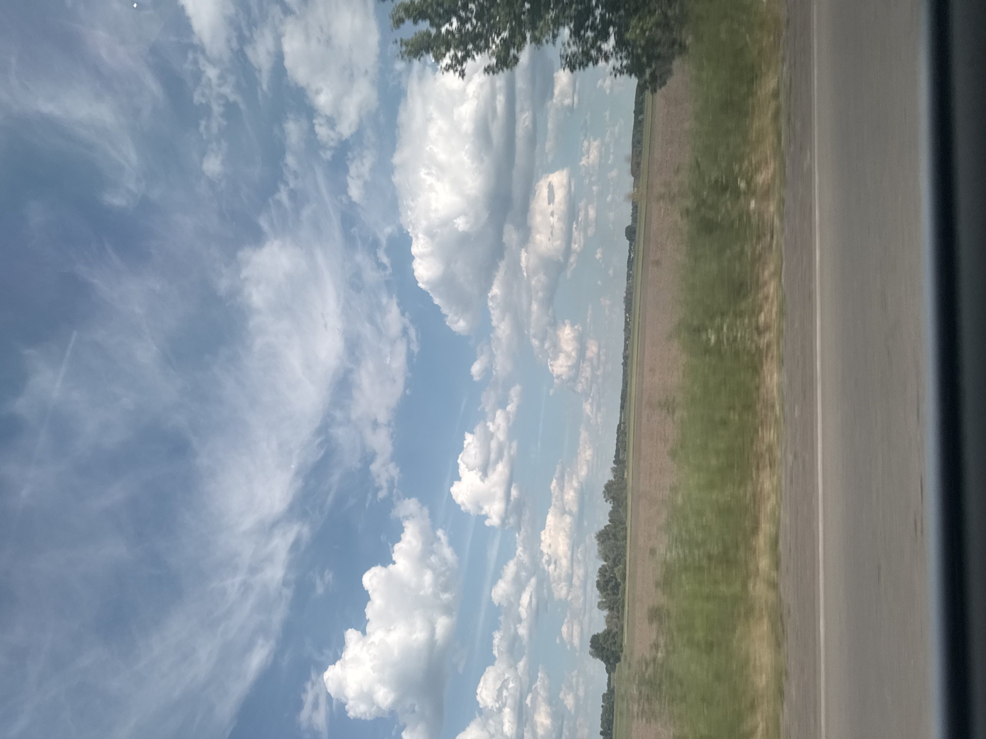 The image depicts a rural landscape near Markivka, Ukraine, observed from a moving vehicle on a bright, partly cloudy day. The sky dominates the upper two-thirds of the frame, showcasing a light blue hue with a mix of cloud formations: wispy, elongated cirrus-type clouds in the upper regions, and distinct, fluffy white cumulus clouds scattered across the mid-sky. A subtle bright spot is present in the upper right sky, possibly the sun obscured by thin cloud or a reflection.
Below the sky, the horizon features an expanse of flat, agricultural fields, primarily appearing as bare, brown soil, interspersed with patches of green vegetation or distant crops. In the mid-distance, several irregular clusters or lines of dark green deciduous trees are visible, marking boundaries or small forested areas. The mid-ground contains a horizontal band of dense, light green vegetation, likely a roadside verge, exhibiting some motion blur.
The immediate foreground reveals a section of a grey asphalt road with a faint white line along its edge, indicating the vantage point is from inside a vehicle traveling on this road. A dark, out-of-focus strip at the very bottom suggests a dashboard or window frame. On the far right, a portion of a tree, characterized by green foliage, is heavily blurred horizontally, consistent with the motion of the viewing vehicle. No people or animals are visible in the scene.