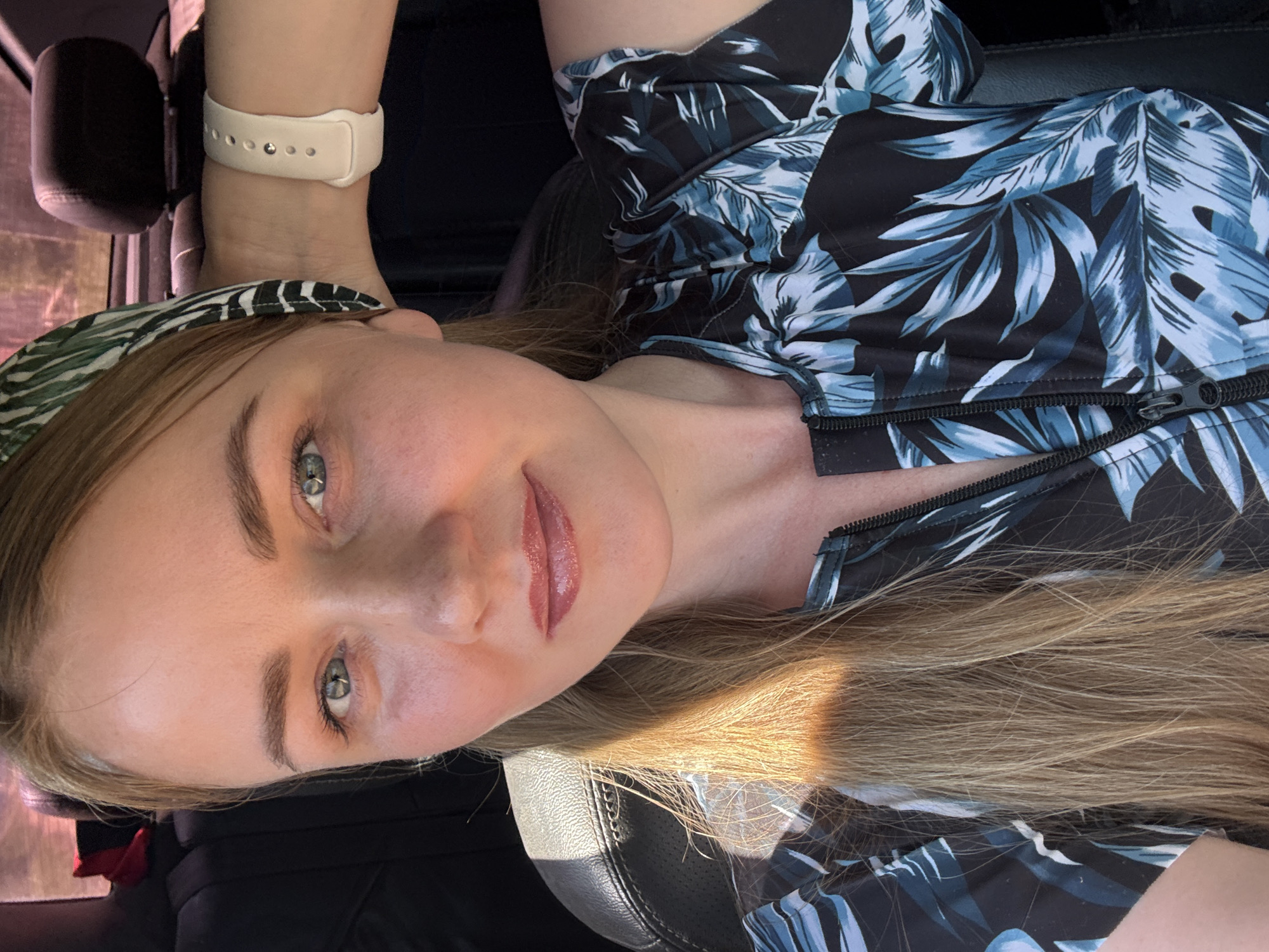 A woman is in the interior of a car, likely sitting in the driver's seat based on the vantage point. She is wearing a black and blue floral pattern top and a headband. She has light-colored hair and is wearing a watch on her left wrist. The location, Chaikovsky, Russia, is not visually discernible from the image.