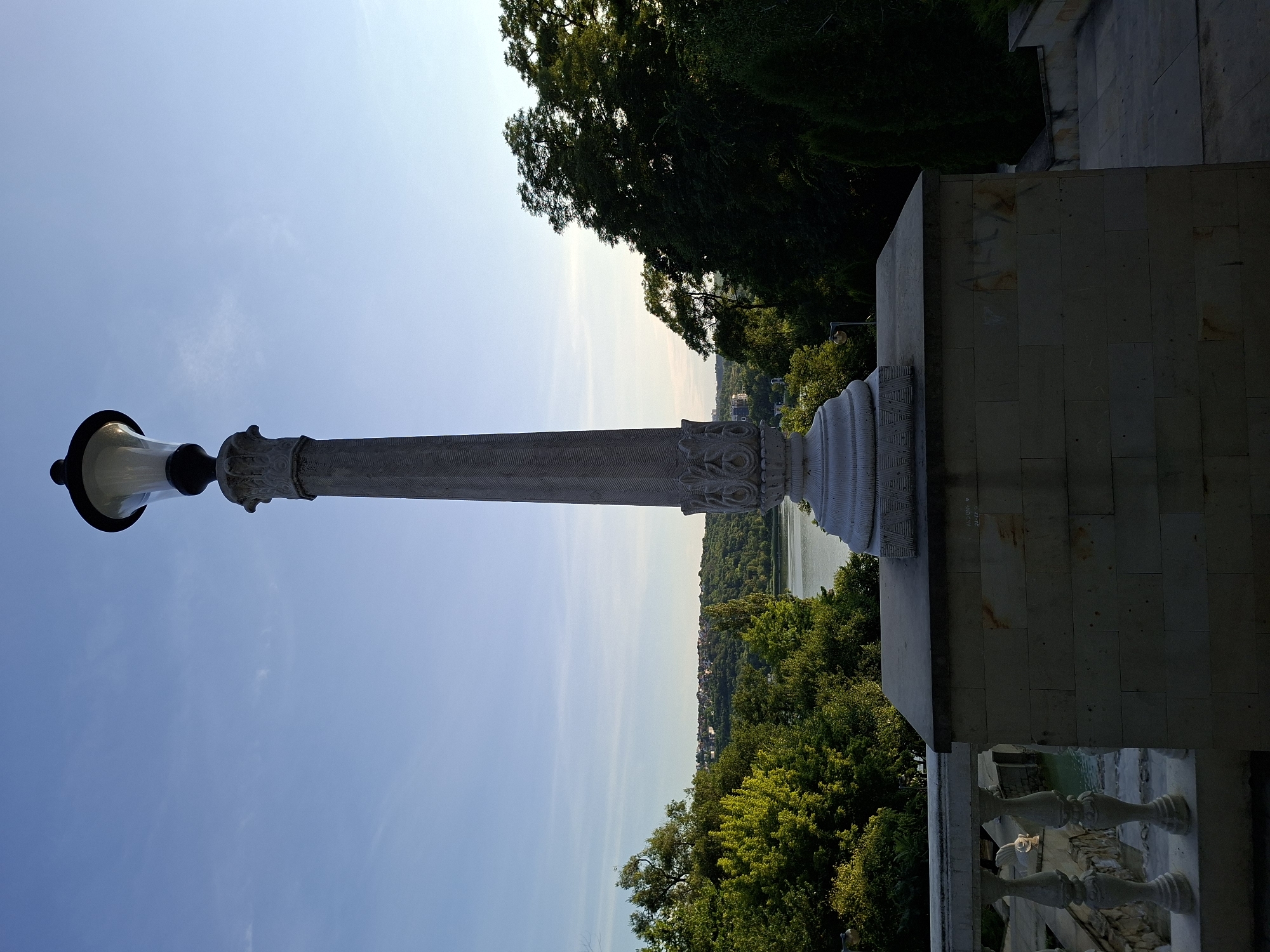 The image depicts a decorative lamppost standing on a stone platform in Chișinău, Moldova. The lamppost is made of stone with ornate carvings. Behind the lamppost, a body of water, likely the Valea Morilor Lake, is visible, surrounded by lush green trees and a distant cityscape. The sky is clear blue with a few wispy clouds. The scene appears to be a park or a scenic overlook, common in Chișinău's recreational areas.