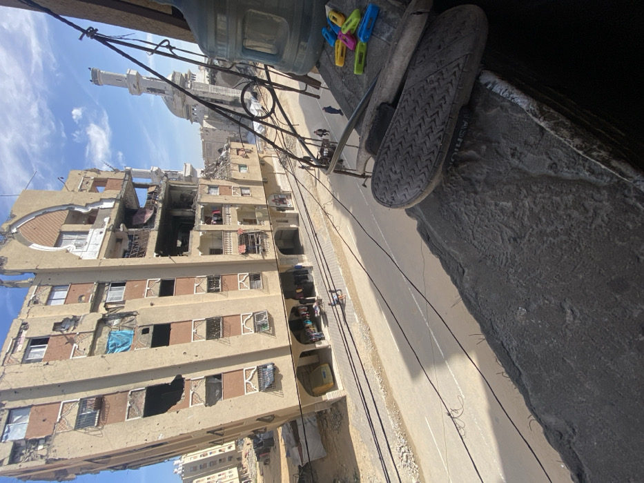 The image captures an urban scene in Khan Yunis, Palestinian Territory, from an elevated perspective looking down onto a street. In the immediate foreground, a dark gray concrete ledge features a pair of dark-colored sandals/slides, one with "FASHION" text visible on its side, and a collection of colorful plastic clothespins (yellow, green, blue, pink, orange). A large, clear plastic water cooler bottle is partially visible to the far right.
The midground is dominated by a paved street and an adjacent sidewalk. Overhead wires are visible, casting shadows across the road. On the left side of the street, a multi-story building, primarily beige and dark brown, displays extensive structural damage, including numerous large holes, missing sections, and exposed interior rooms. Clothes are visible hanging from some balconies. A small yellow vehicle is parked beneath an arched entrance on the ground floor. Further down the street, on the right side, a mosque with a tall minaret and a large dome is visible. Several individuals are walking on the sidewalk and street below, with two figures prominent near the center. Additional damaged or partially constructed buildings are visible in the background on the far left. The sky is blue with scattered white clouds.