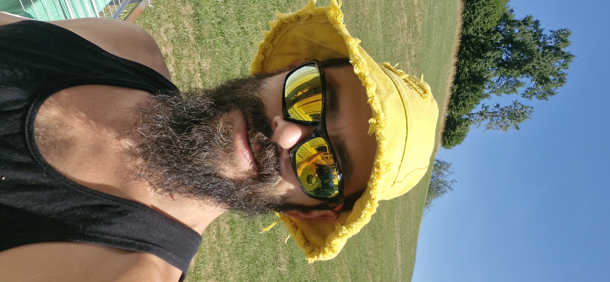 A close-up, low-angle selfie shows a man with a full beard and mustache wearing a yellow bucket hat and black sunglasses outdoors on a sunny day. The man's face is the primary focus, with his sunglasses reflecting a yellow structure and a green bucket. Behind him, a grassy hillside slopes upwards, dotted with trees and shrubs. The sky is clear blue. The location is the village of Wattenwil, Switzerland, specifically on the hill above the village, with a view towards the south-east. The man appears to be in a relaxed, recreational setting.