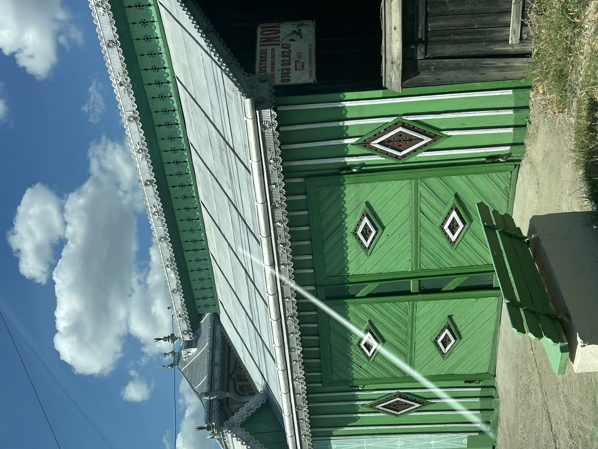 In the city of Băișești, Romania, a vibrant green wooden building with white trim stands under a bright blue sky with scattered white clouds. The building features a gabled roof with intricate white decorative edging along the eaves. The facade is constructed of vertical wooden planks painted bright green, interspersed with white vertical stripes. A large set of double doors, also painted green, dominates the center of the facade, each door decorated with a diamond-shaped panel containing a brown diamond with a white border. To the right of the doors, a sign with red lettering on a white background is affixed to the building. The sign reads "LUSTRADE INOX" and displays a phone number "0743 02.02.47" along with some smaller text that is difficult to discern. To the left of the doors, a white beam of light, possibly from the sun, cuts diagonally across the image. In the foreground, a simple green wooden bench sits on a concrete base, facing away from the building. The ground in front of the building appears to be a mix of concrete and dirt, with patches of dry grass. To the far right, a section of a dark wooden fence is visible. The overall impression is one of a traditional Romanian building, likely a shop or residence, with decorative architectural elements.
