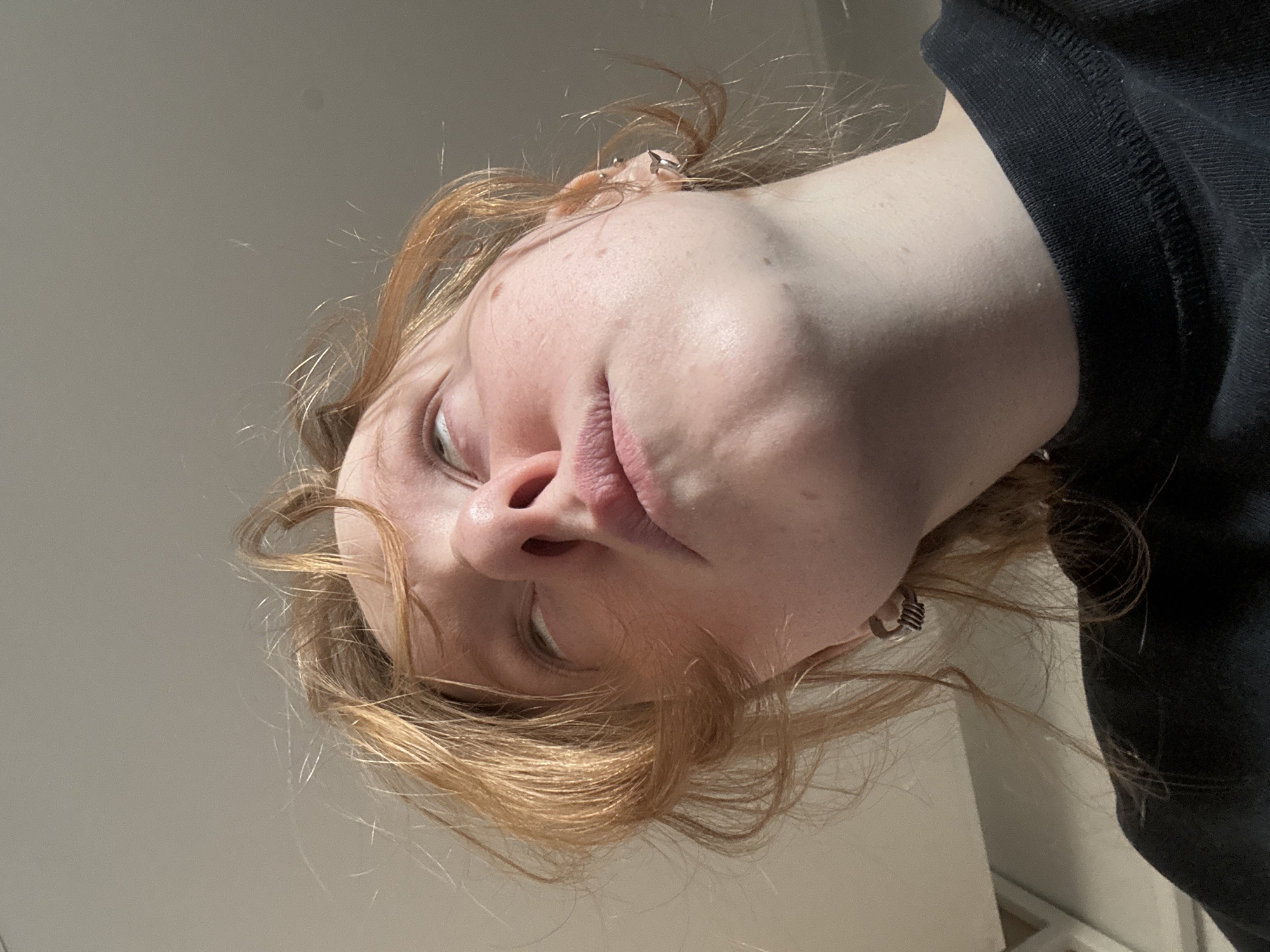 A low-angle photograph depicts a person with fair skin and reddish-blonde hair. The person is wearing a dark-colored top. There are piercings visible in the ear. The location is not discernible as Perm, Russia. The environment appears to be indoors, given the presence of a wall in the background.
