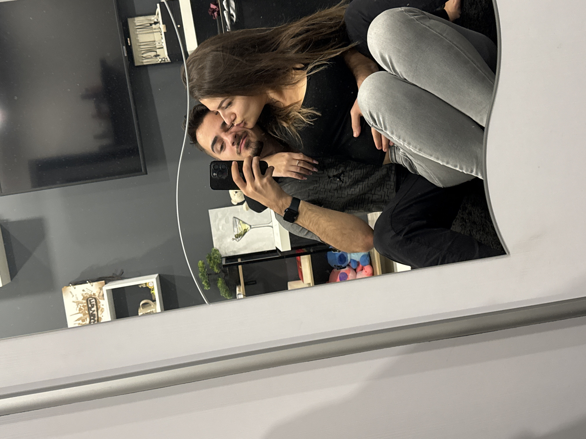 A mirrored selfie shows a couple. A woman with long brown hair wearing a black shirt kisses a man with a beard and dark hair on the cheek. The man is holding a black smartphone and wearing a watch on his left wrist. The woman has a ring on her left hand. They are seated. The background includes a wall-mounted TV, shelves with decor, and a piece of art depicting a martini glass. The exact location in Sofia, Bulgaria cannot be determined from the image.