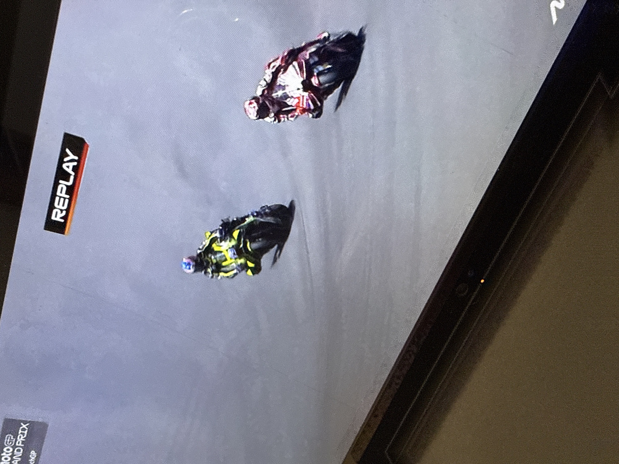 The image shows a replay of a motorcycle race on a screen. Two motorcycles are visible, one red and white and the other yellow and black, racing on a gray track. The red and white motorcycle is in the lead, with the yellow and black motorcycle following closely behind. The "REPLAY" logo is visible on the left side of the screen, indicating that the footage is a replay. The "MotoGP GRAND PRIX" text is also visible at the bottom left of the screen. The scene appears to be a sporting event, likely a professional motorcycle race. There are no clear indicators of time of day or weather conditions. The image is a photograph of a screen, and the overall setting is indoors, as indicated by the surrounding dark frame and the textured background on the right.