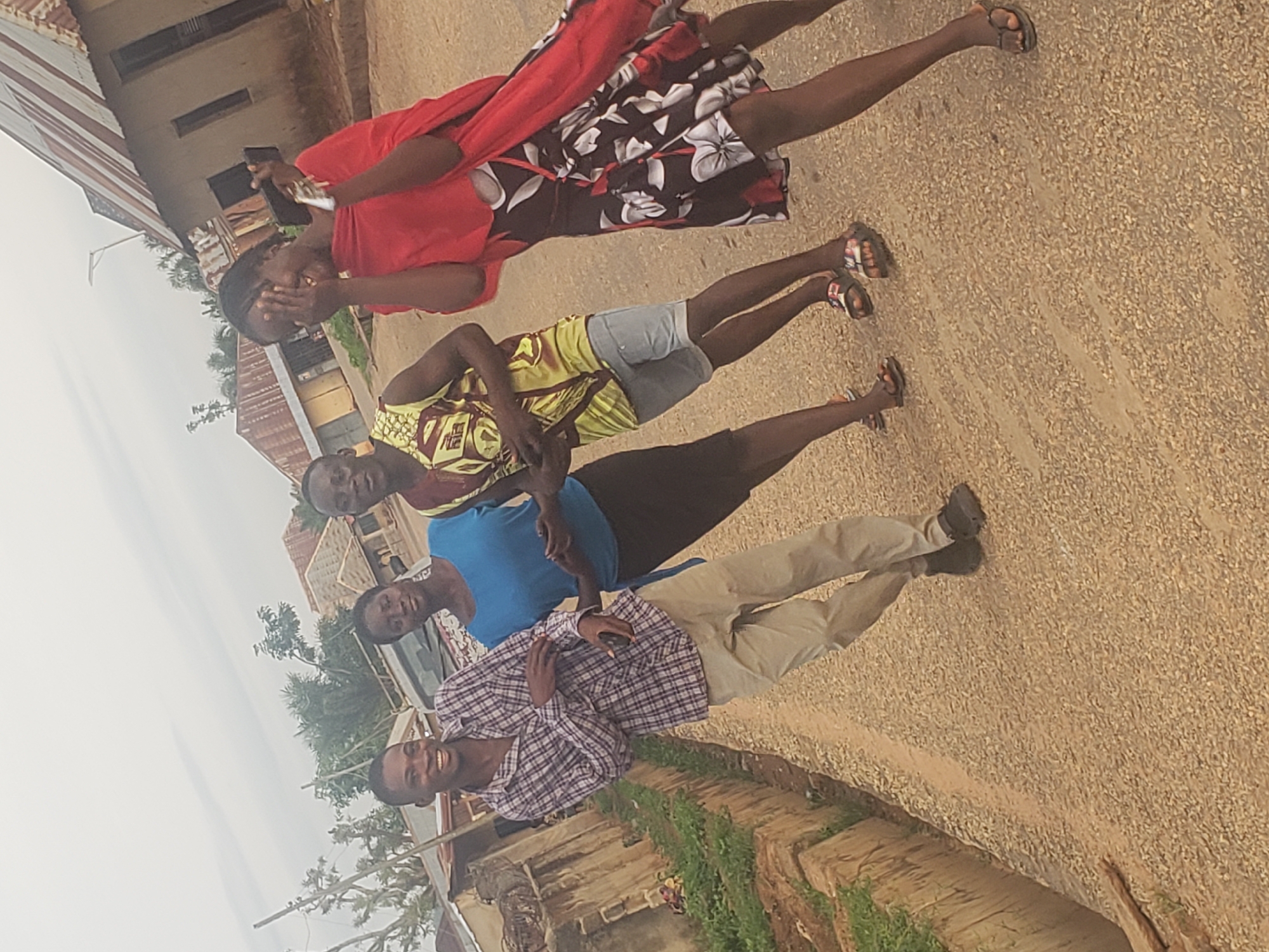 The image depicts four young individuals outdoors on a dirt road, likely in the city of Gumel, Nigeria. The individuals are positioned in a line, with their bodies angled towards the viewer. The person on the far left is a male wearing a checkered shirt and light-colored pants. Next to him is a female in a blue top and dark skirt. To her right is a male in a sleeveless patterned shirt and shorts. The person on the far right is a female wearing a red wrap over a floral print dress. She appears to be holding a phone in her right hand and covering her face with her left, possibly in a gesture of surprise or shyness. The background shows a collection of buildings with corrugated metal roofs, characteristic of residential structures in the region. The ground is a mix of dirt and small gravel. The sky is overcast, suggesting a cloudy day.